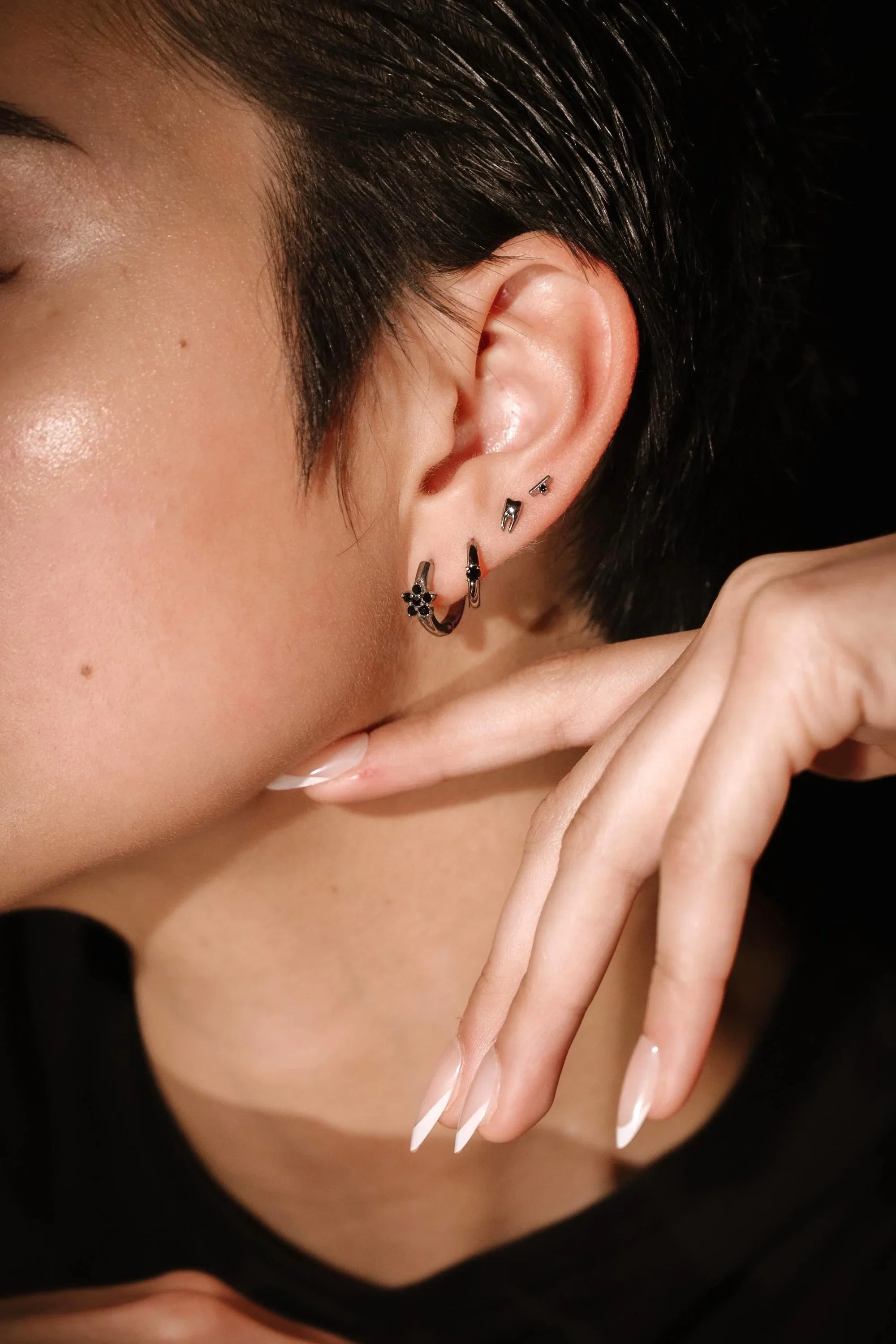 Close-up of an ear with several silver and black earrings and a hand touching the face, against a dark background