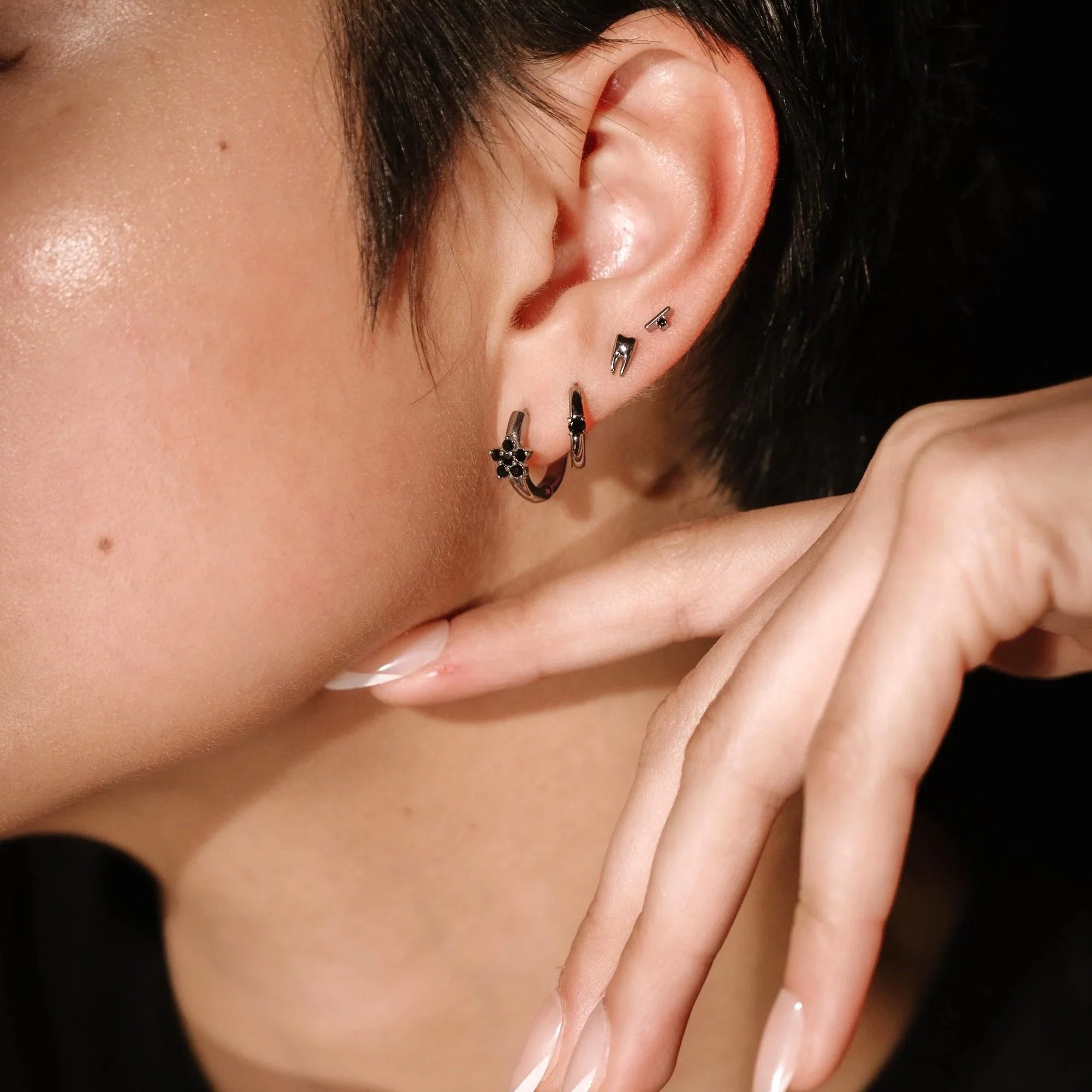 Close-up of an ear with several silver and black earrings and a hand touching the face, against a dark background
