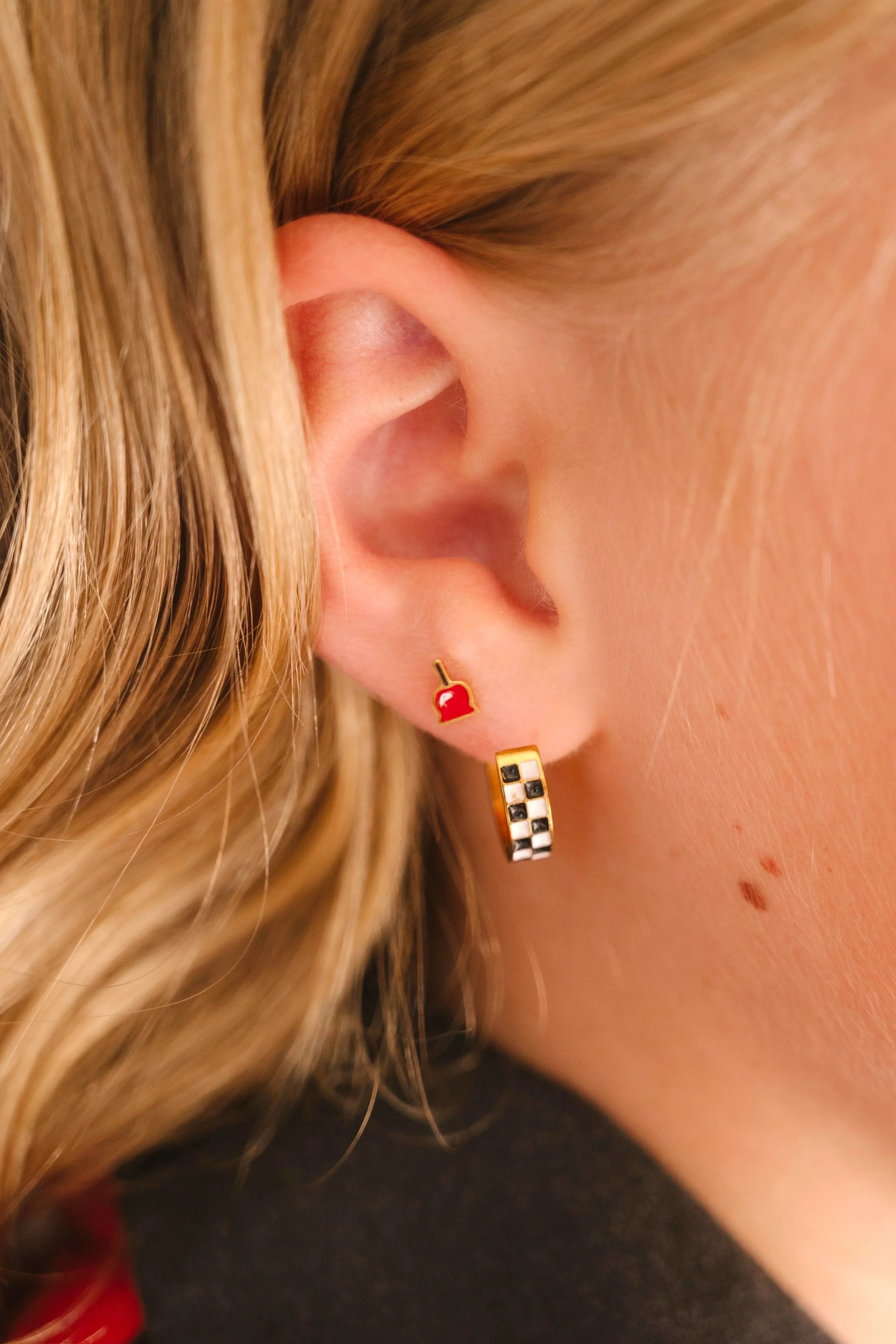 Close-up of a person wearing a checkered hoop earring in a first lobe piercing and a bright red enamel stud shaped like a candied apple in a second lobe piercing.