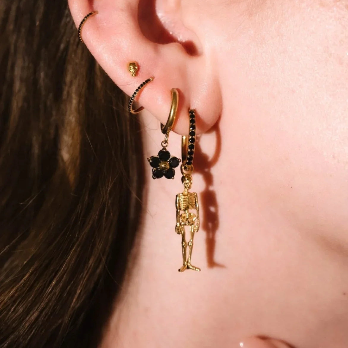 Close up of an ear with black and gold flat backs and hoops, with a detailed skeleton charm dangling from one.