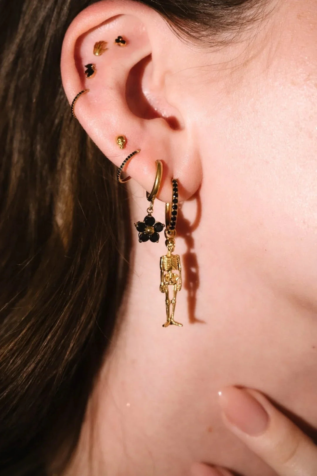 Close up of an ear with black and gold flat backs and hoops, with a detailed skeleton charm dangling from one.
