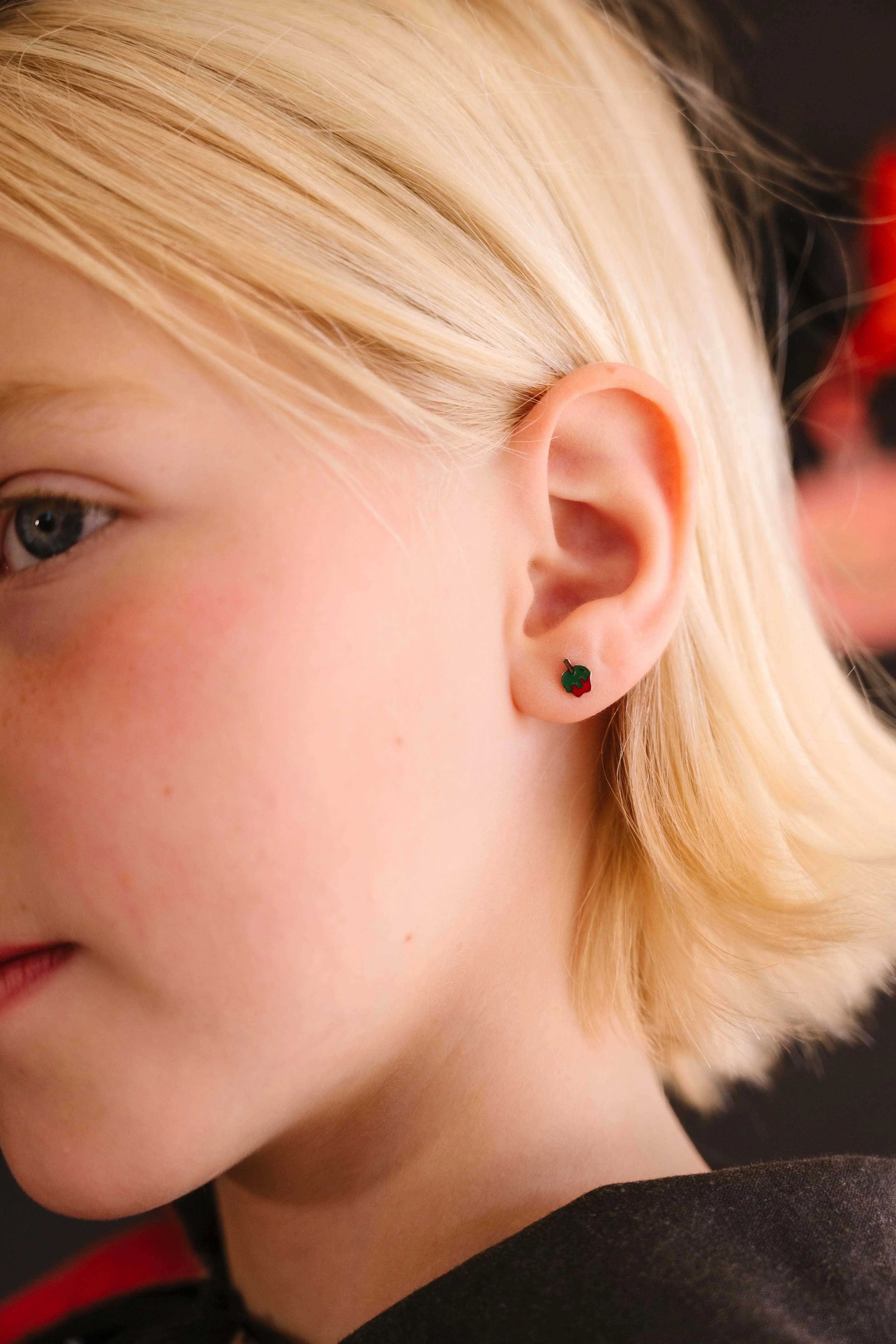 Close-up of a person wearing a small green and red enamel earring shaped like an apple dipped in green poison.