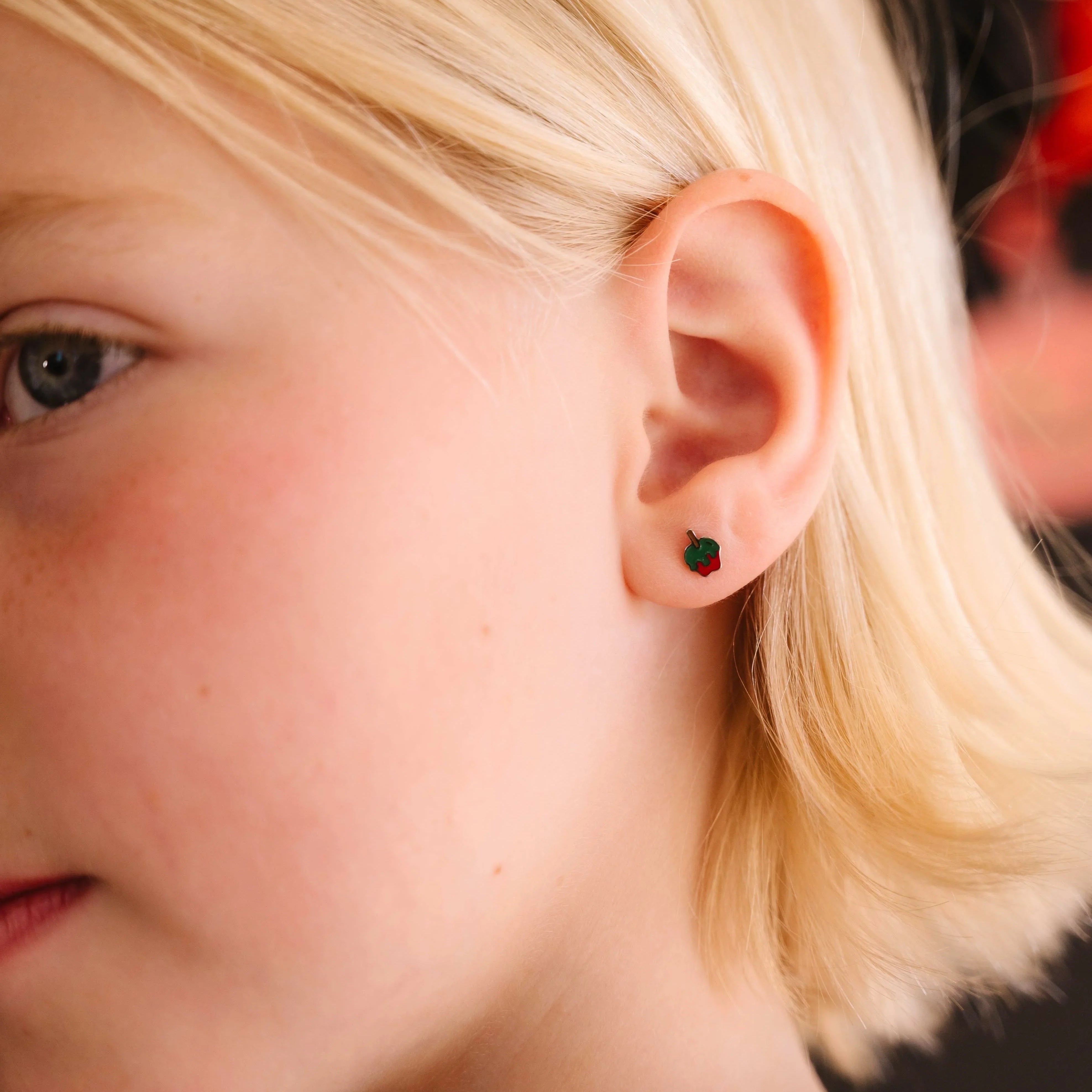 Close-up of a person wearing a small green and red enamel earring shaped like an apple dipped in green poison.
