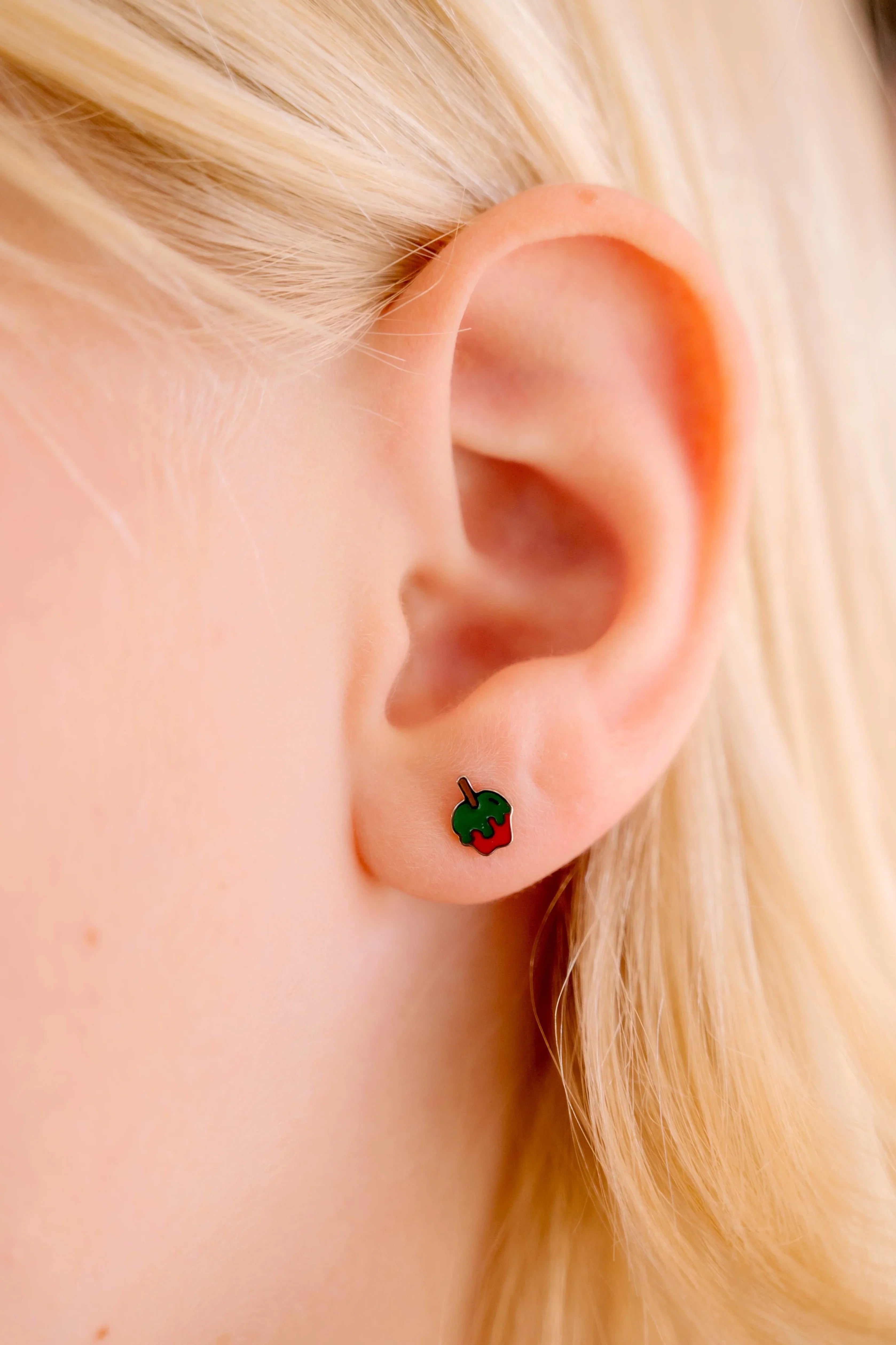 Close-up of an ear wearing a small green and red enamel earring shaped like an apple dipped in green poison.