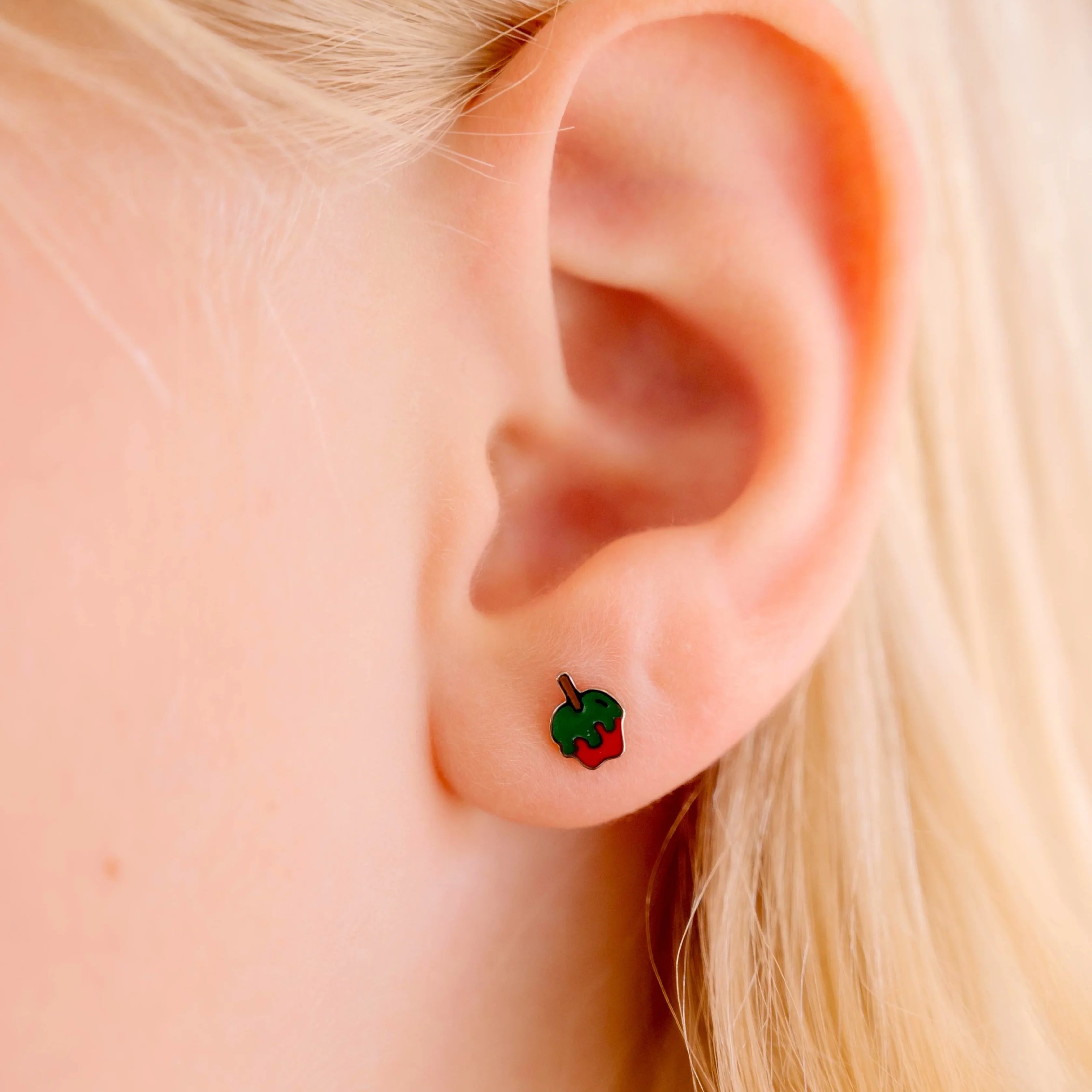 Close-up of an ear wearing a small green and red enamel earring shaped like an apple dipped in green poison.