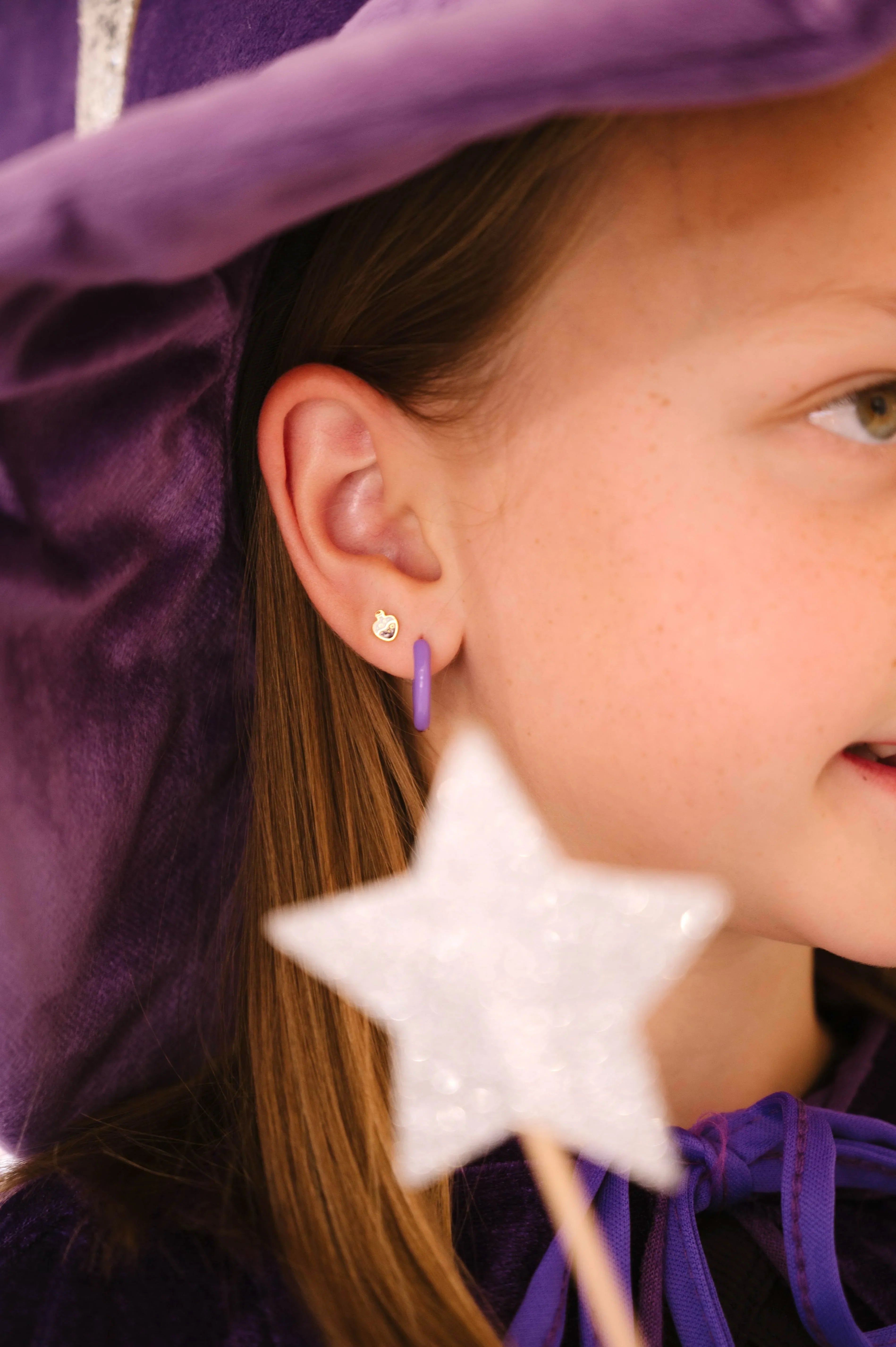 Close-up of a child wearing a purple witch hat and outfit, with a focus on a purple enamel potion bottle-shaped stud earring.