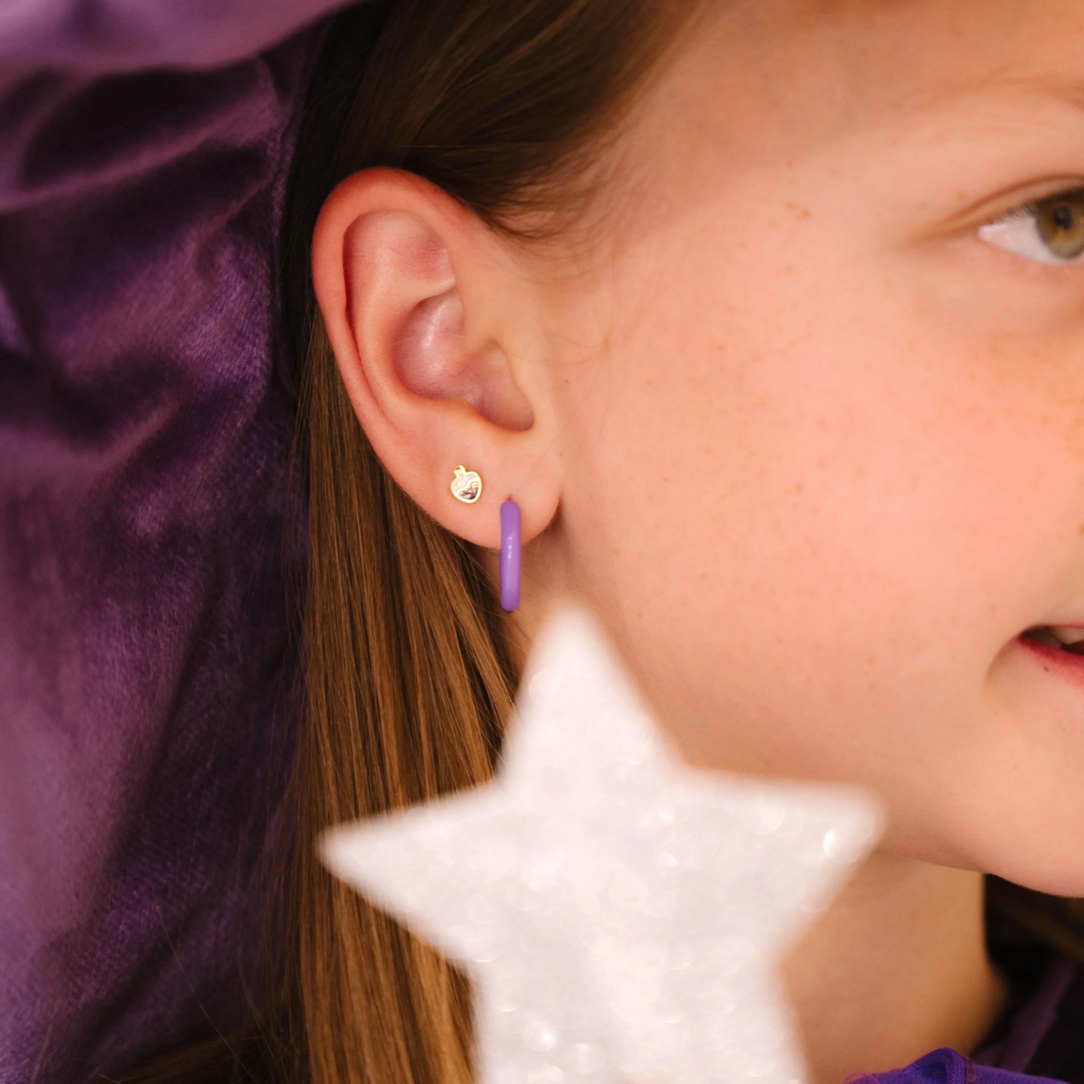 Close-up of a child wearing a purple witch hat and outfit, with a focus on a purple enamel potion bottle-shaped stud earring.