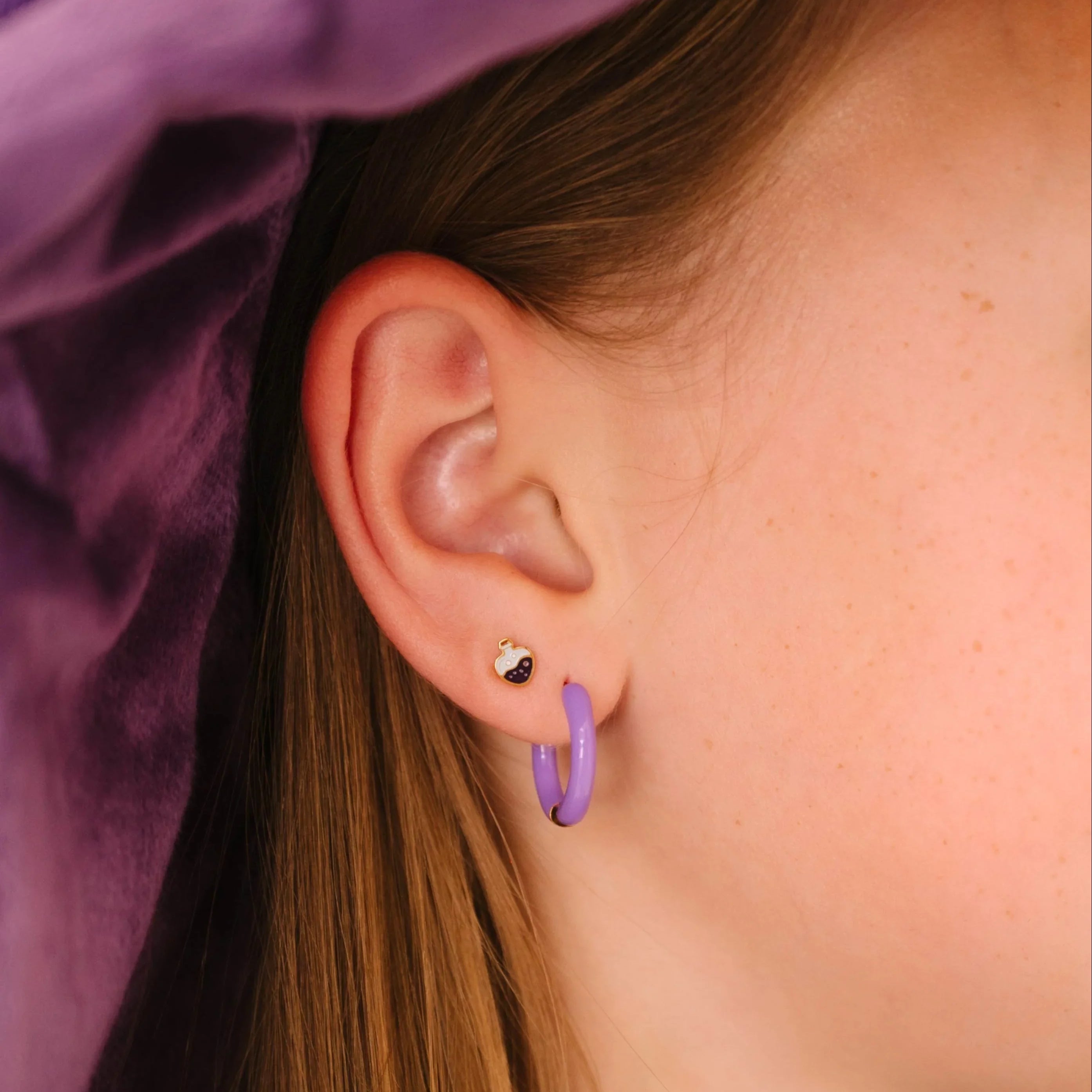 Close-up of a child wearing a purple witch hat and outfit, with a focus on a purple enamel potion bottle-shaped stud earring.