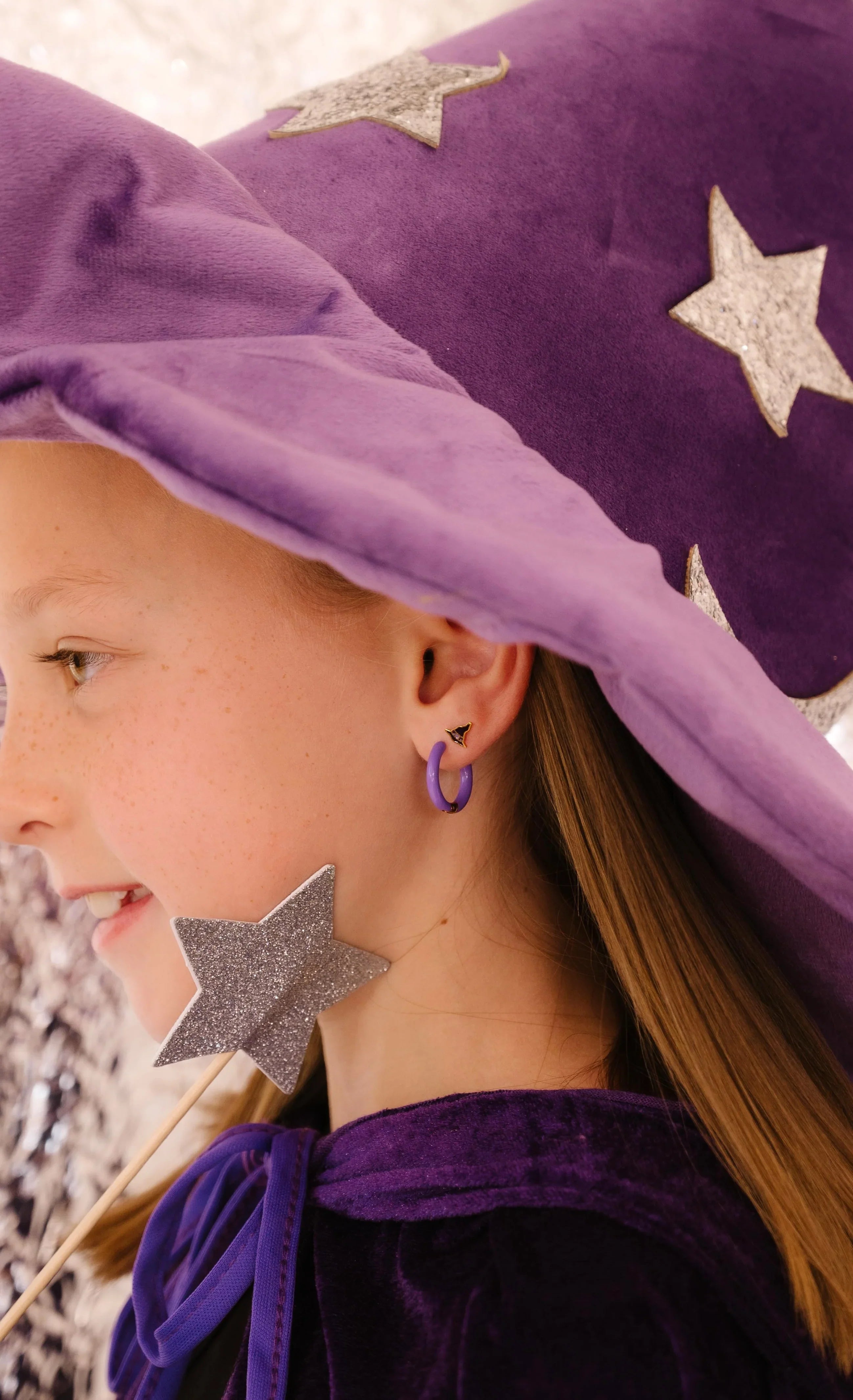 Child wearing a purple witch hat with star decorations and a matching outfit. A focus on the child's jewelry, including a purple hoop earring and a dark purple and gold stud earring, with a blurred background
