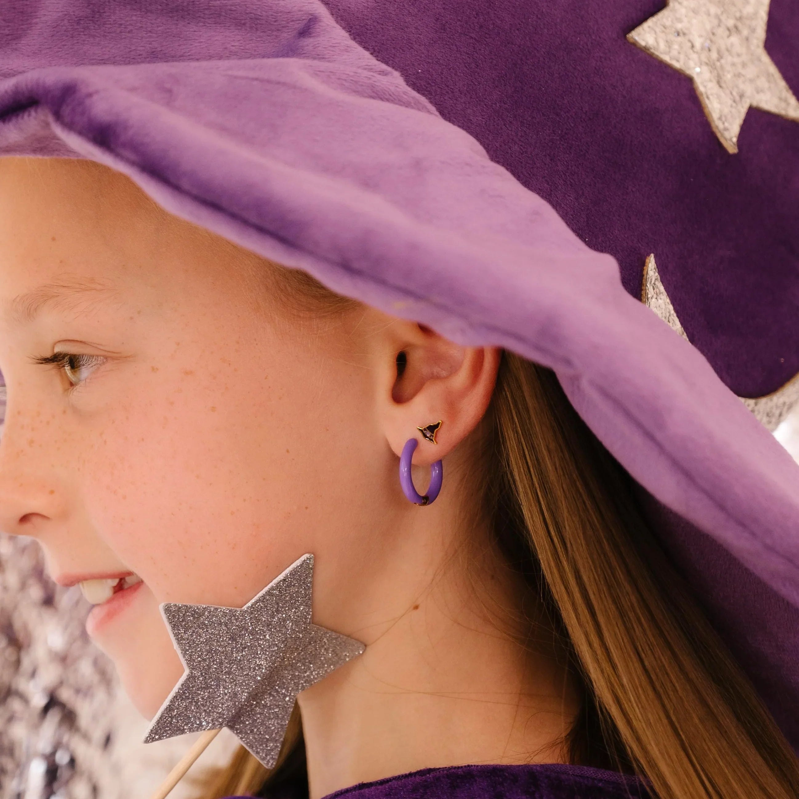 Child wearing a purple witch hat with star decorations and a matching outfit. A focus on the child's jewelry, including a purple hoop earring and a dark purple and gold stud earring, with a blurred background