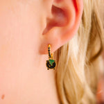 Close-up of an ear wearing a gold earring with green and black enamel charm shaped like a cauldron.