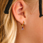Close-up of an ear wearing a gold earring with a purple enamel hoop charm shaped like a witch's hat, with blonde hair and black headband.