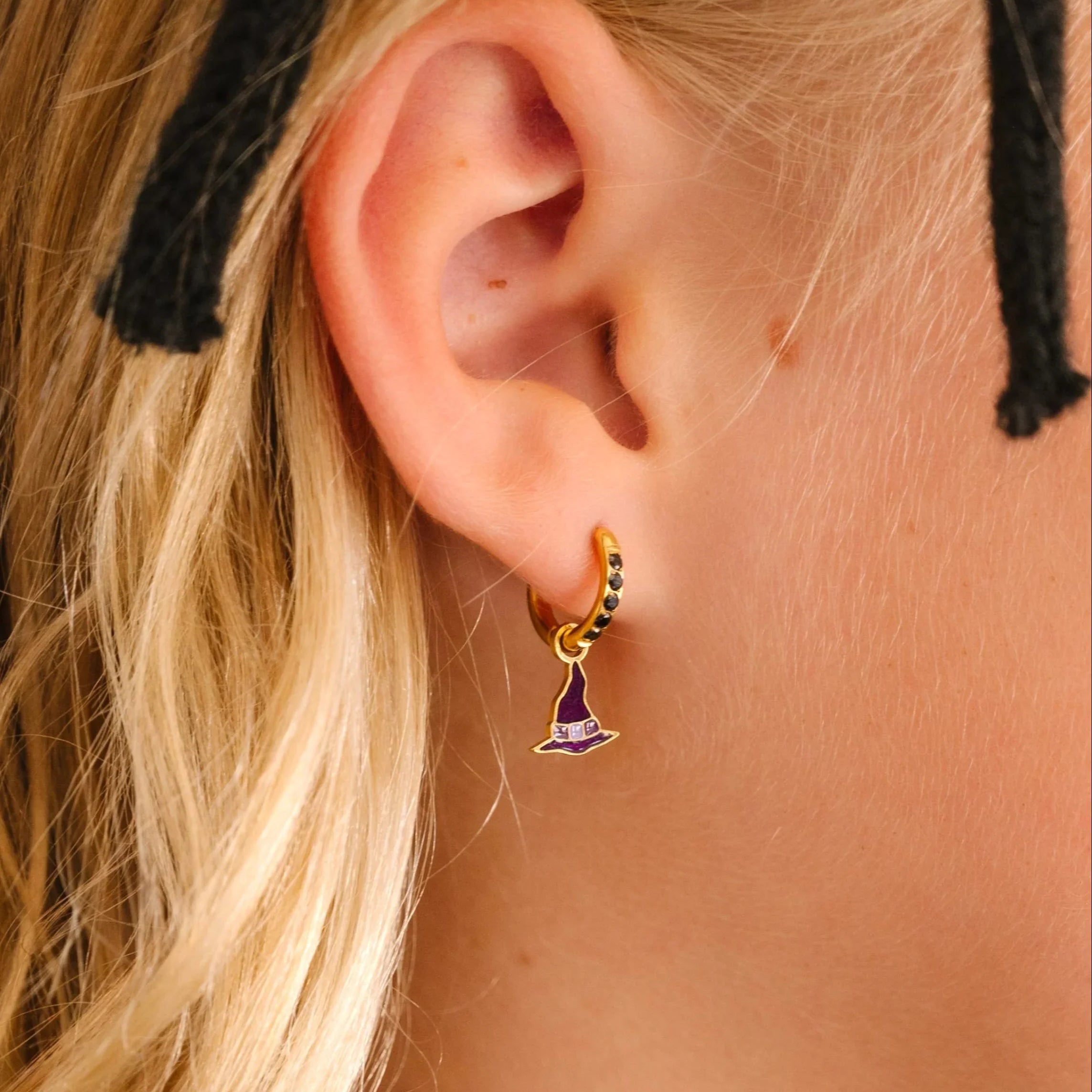 Close-up of an ear wearing a gold earring with a purple enamel hoop charm shaped like a witch's hat, with blonde hair and black headband.