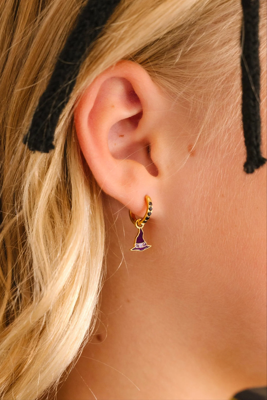 Close-up of an ear wearing a gold earring with a purple enamel hoop charm shaped like a witch's hat, with blonde hair and black headband.