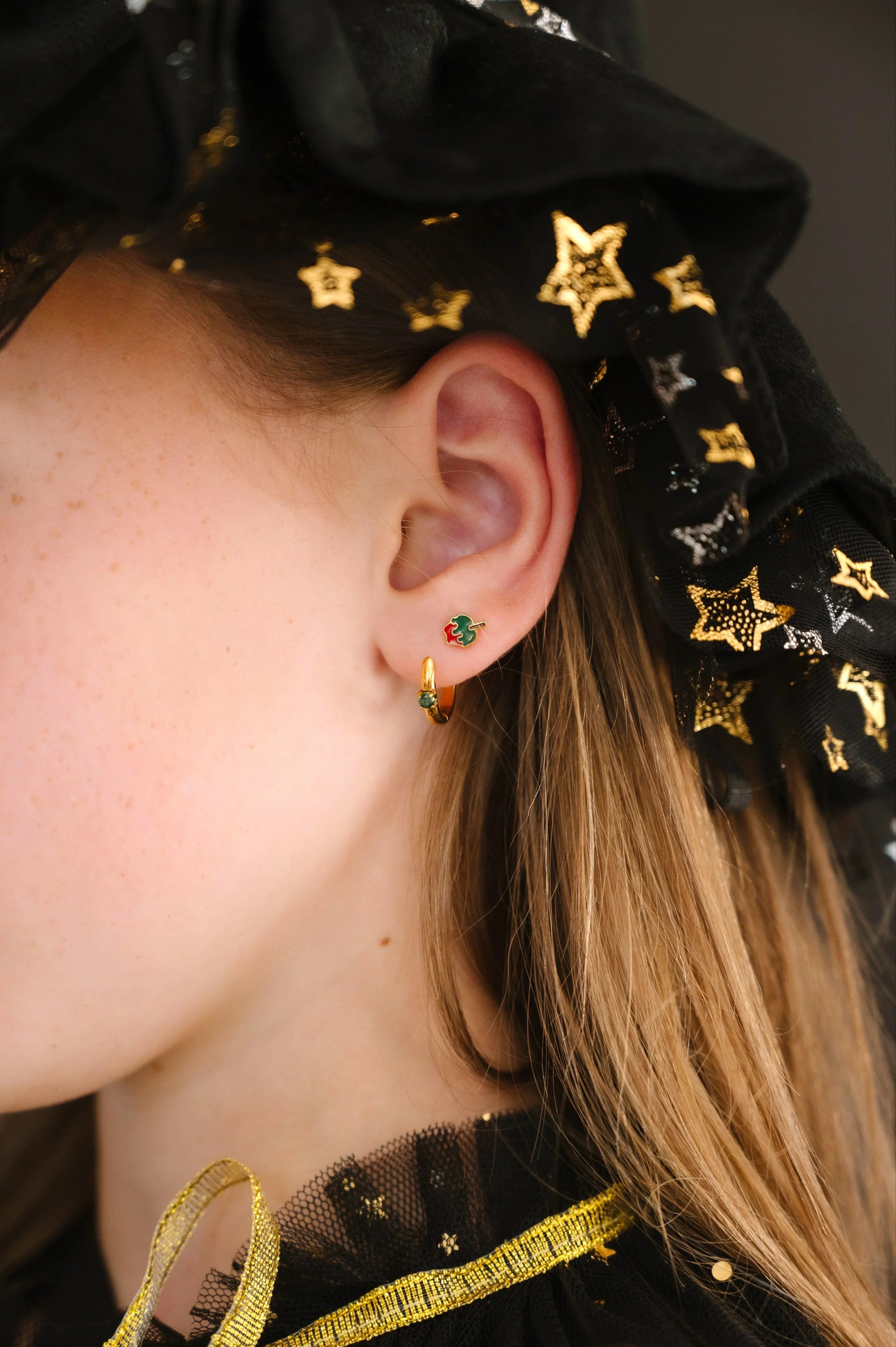 Close-up of an ear wearing gold earrings with green and red enamel, against a dark background with star patterns.