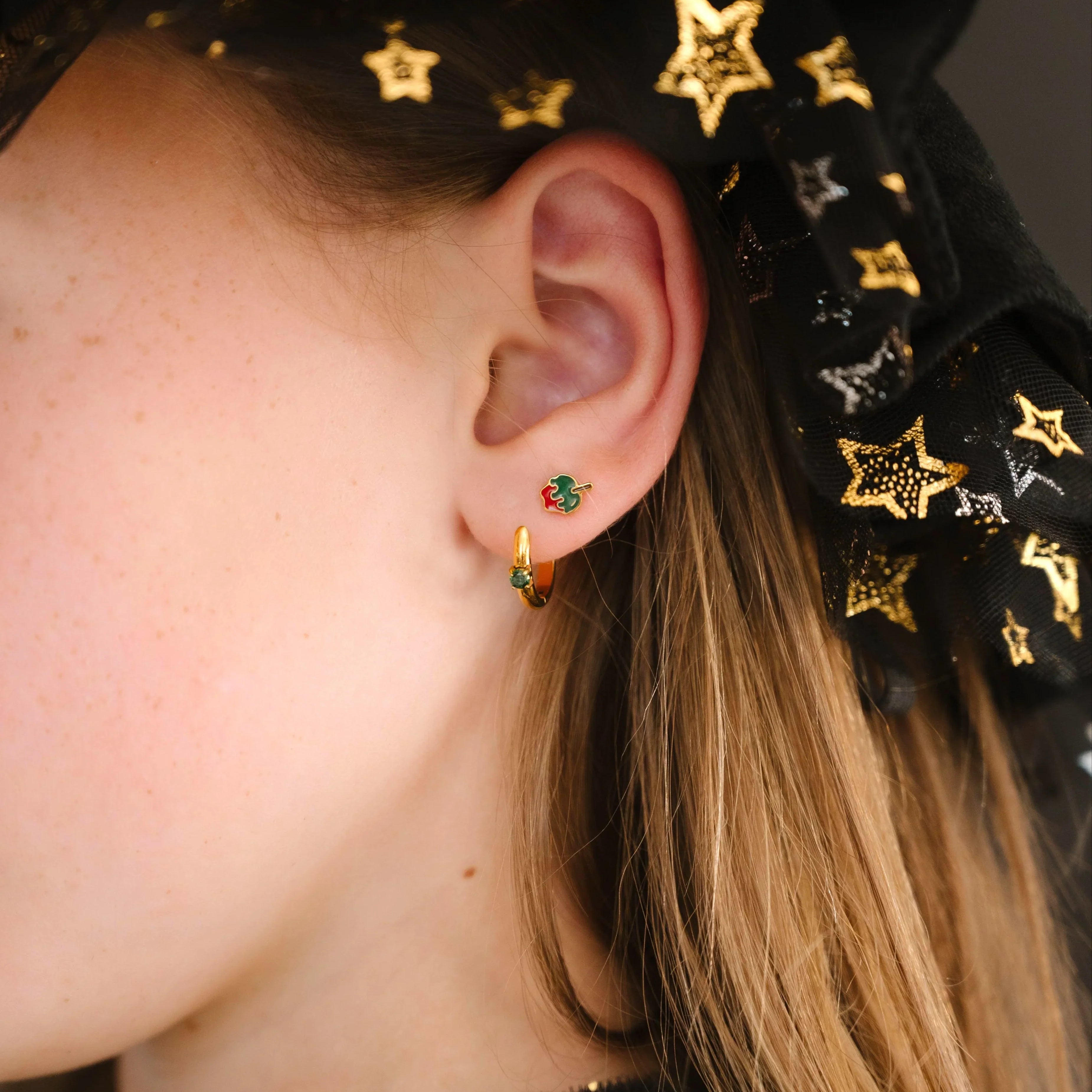 Close-up of an ear wearing gold earrings with green and red enamel, against a dark background with star patterns.