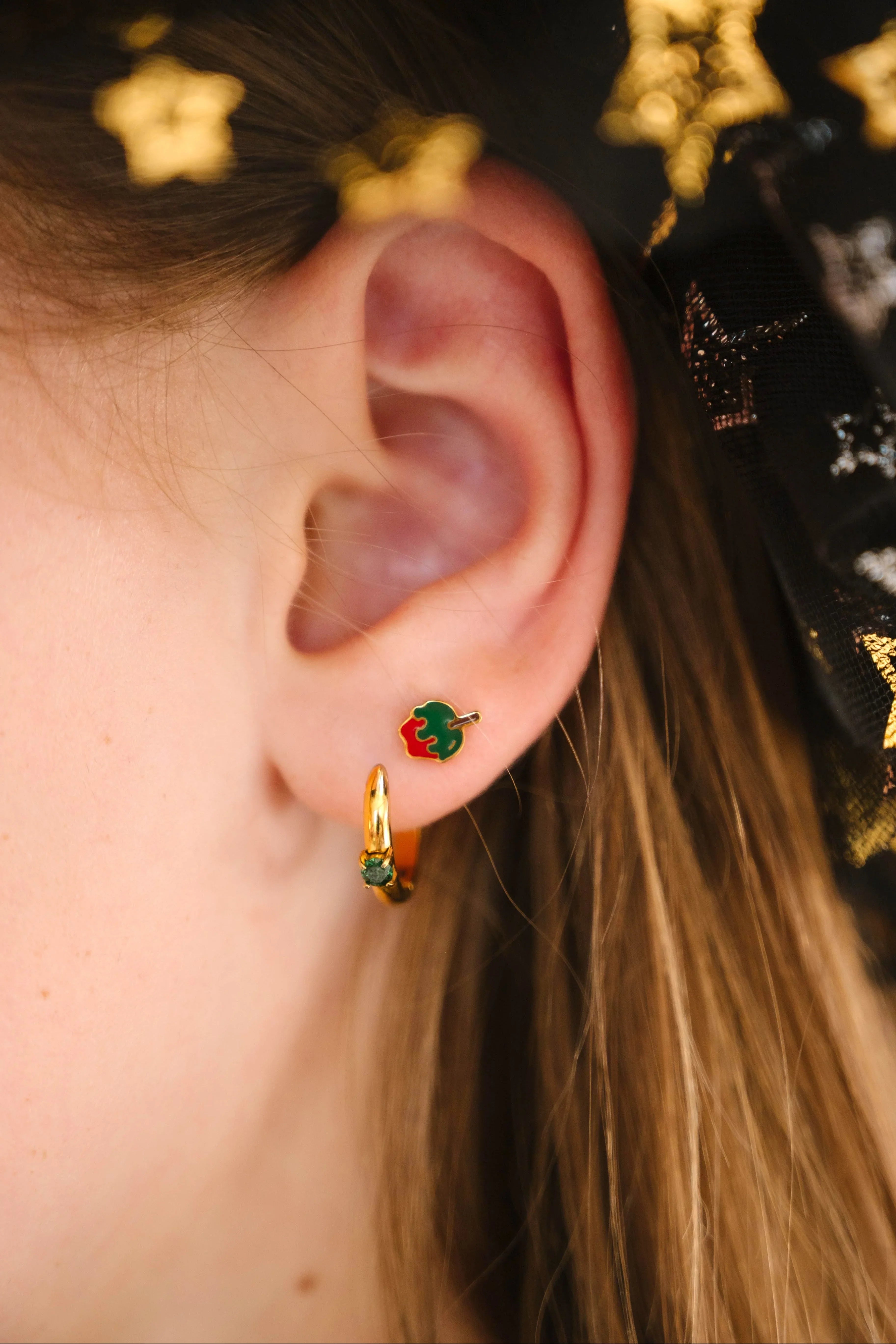 Close-up of an ear wearing gold earrings with green and red enamel, against a dark background with star patterns.