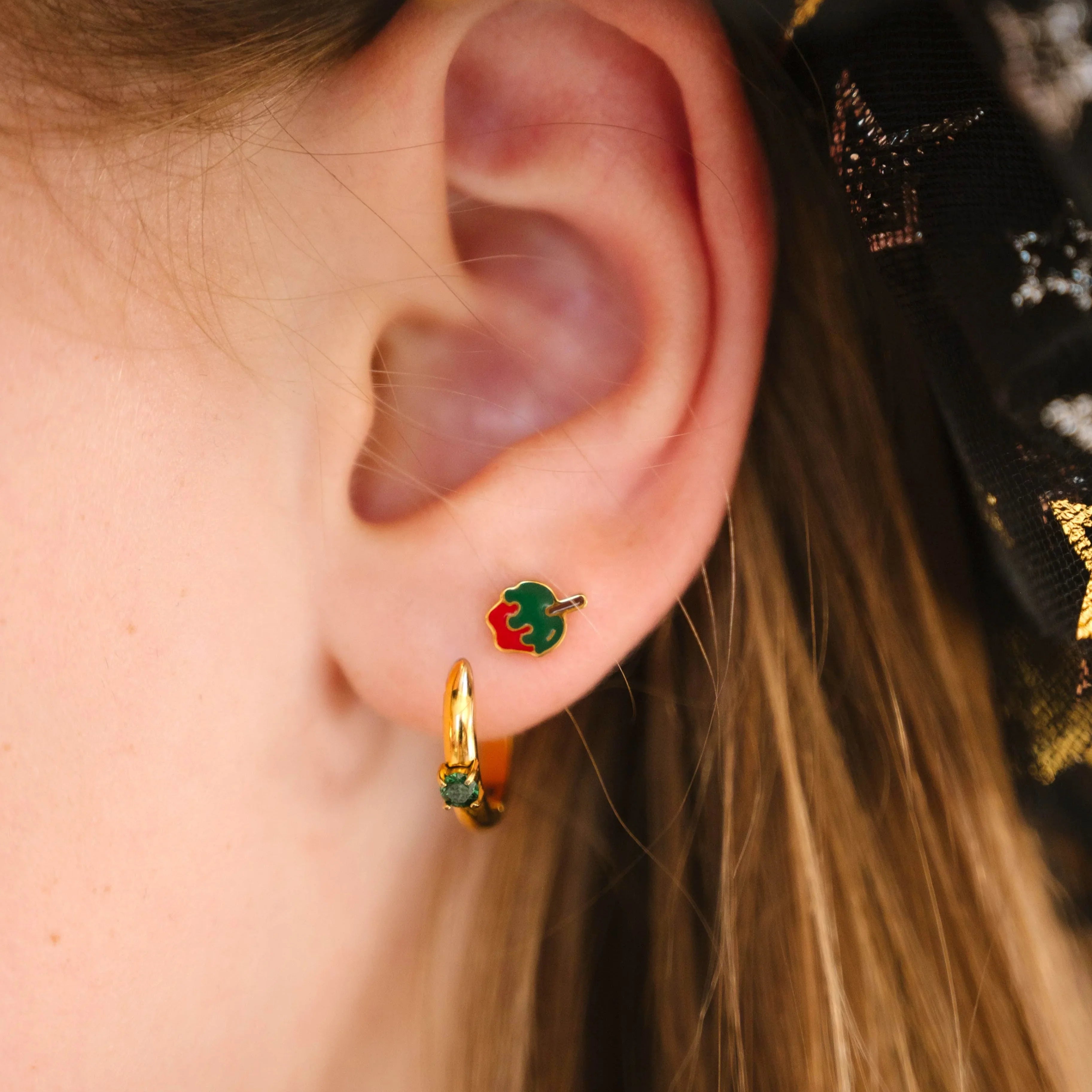 Close-up of an ear wearing gold earrings with green and red enamel, against a dark background with star patterns.