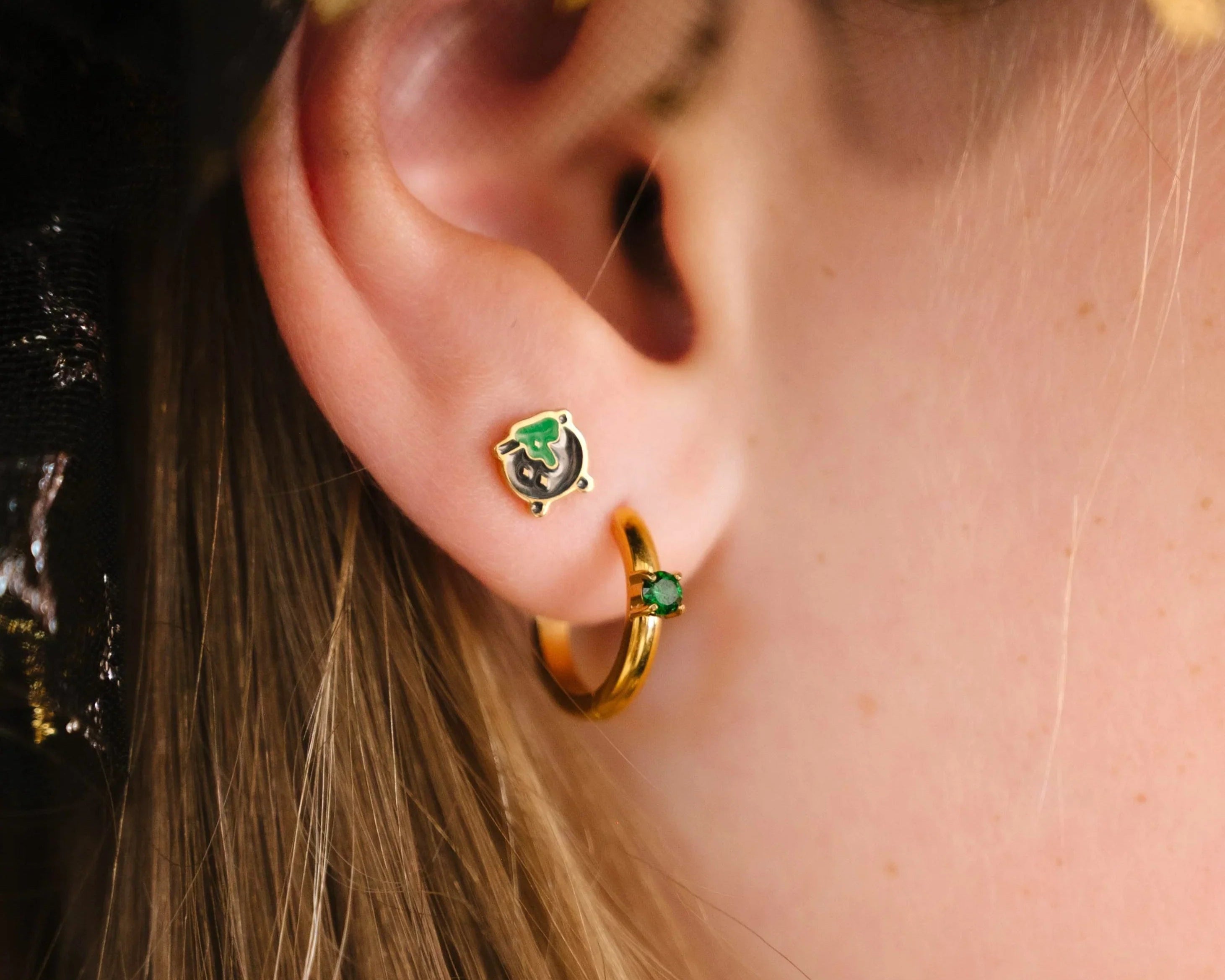 Close-up of an ear wearing gold hoop earrings with green gemstones and a black, green, and gold cauldron-shaped stud.