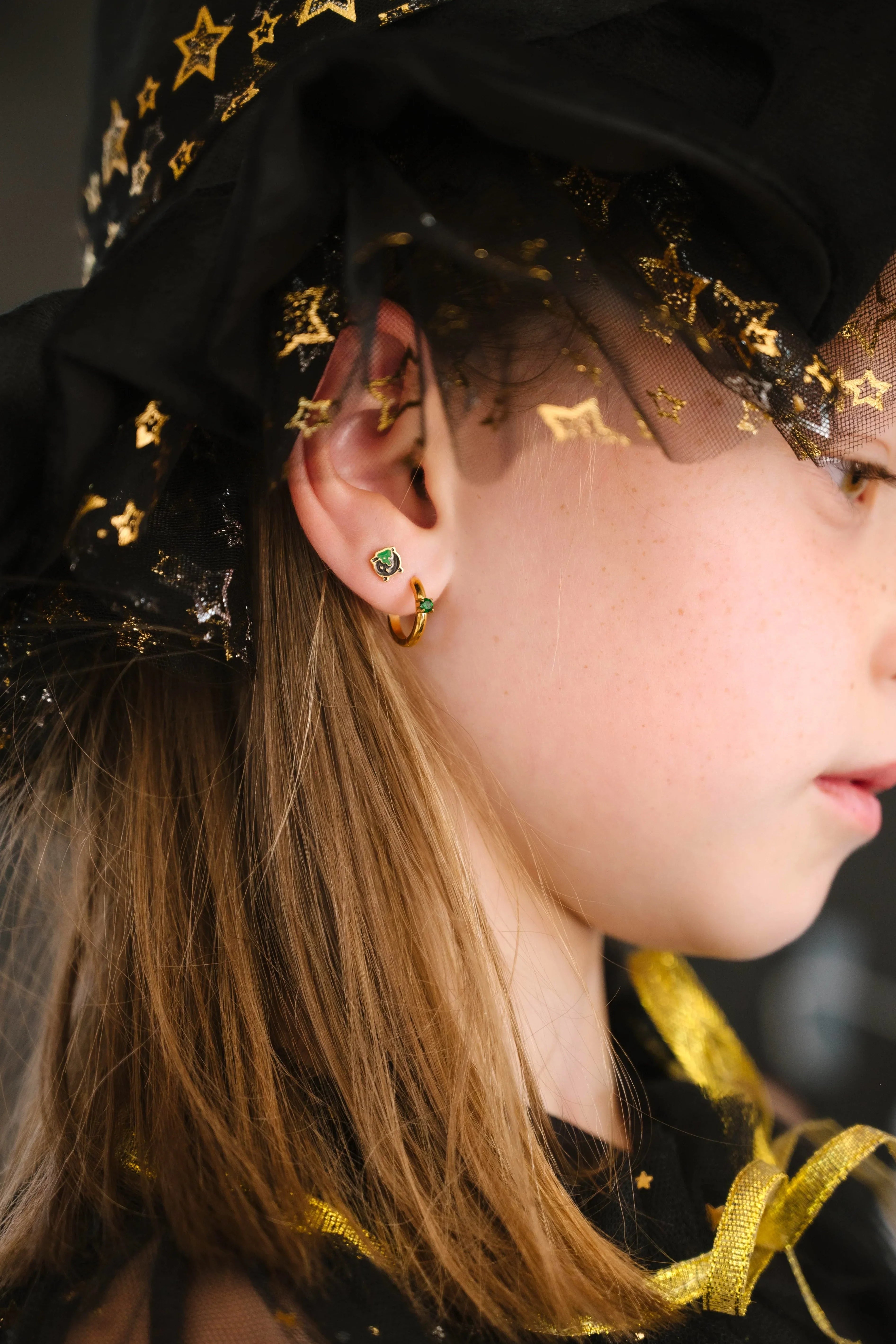 Close-up of an ear wearing gold hoop earrings with green gemstones and a black, green, and gold cauldron-shaped stud.
