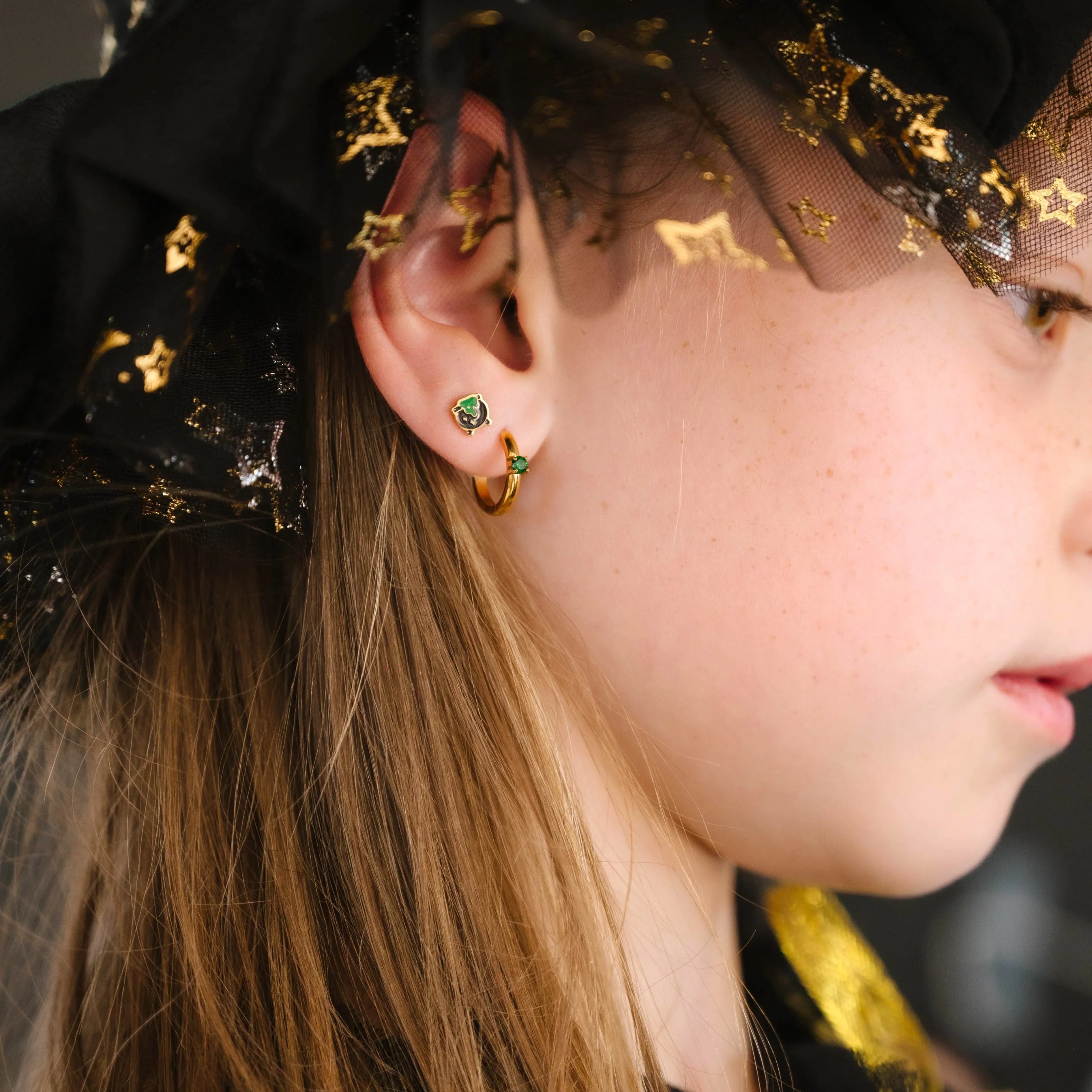Close-up of an ear wearing gold hoop earrings with green gemstones and a black, green, and gold cauldron-shaped stud.