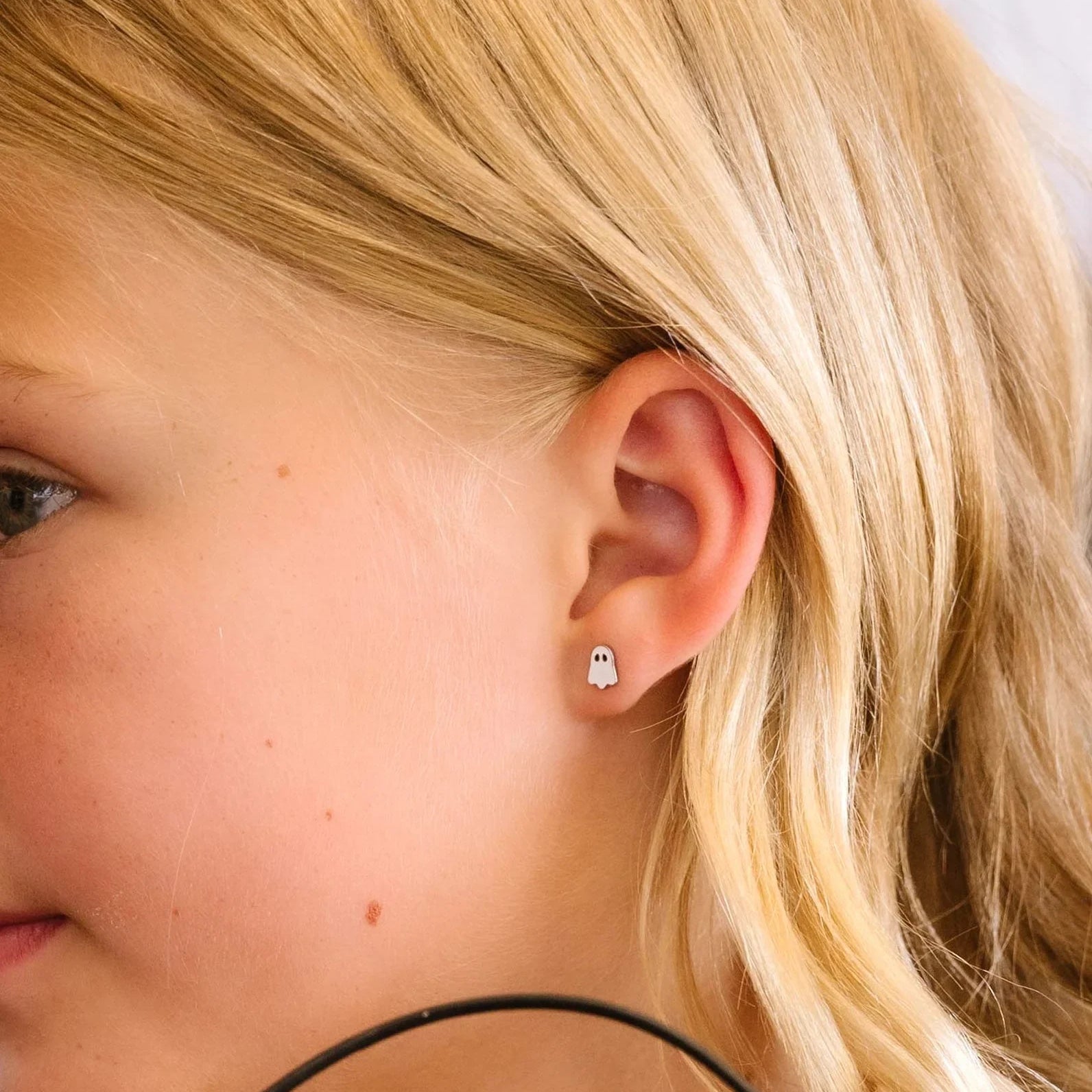 Close-up of a young girl with blonde hair wearing a small ghost-shaped white stud earring.