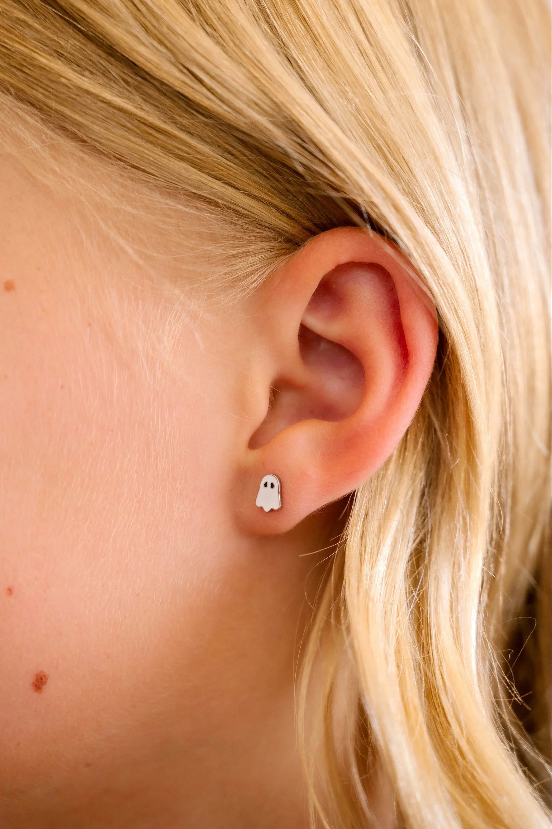Close-up of an ear wearing a small ghost-shaped earring with blonde hair.