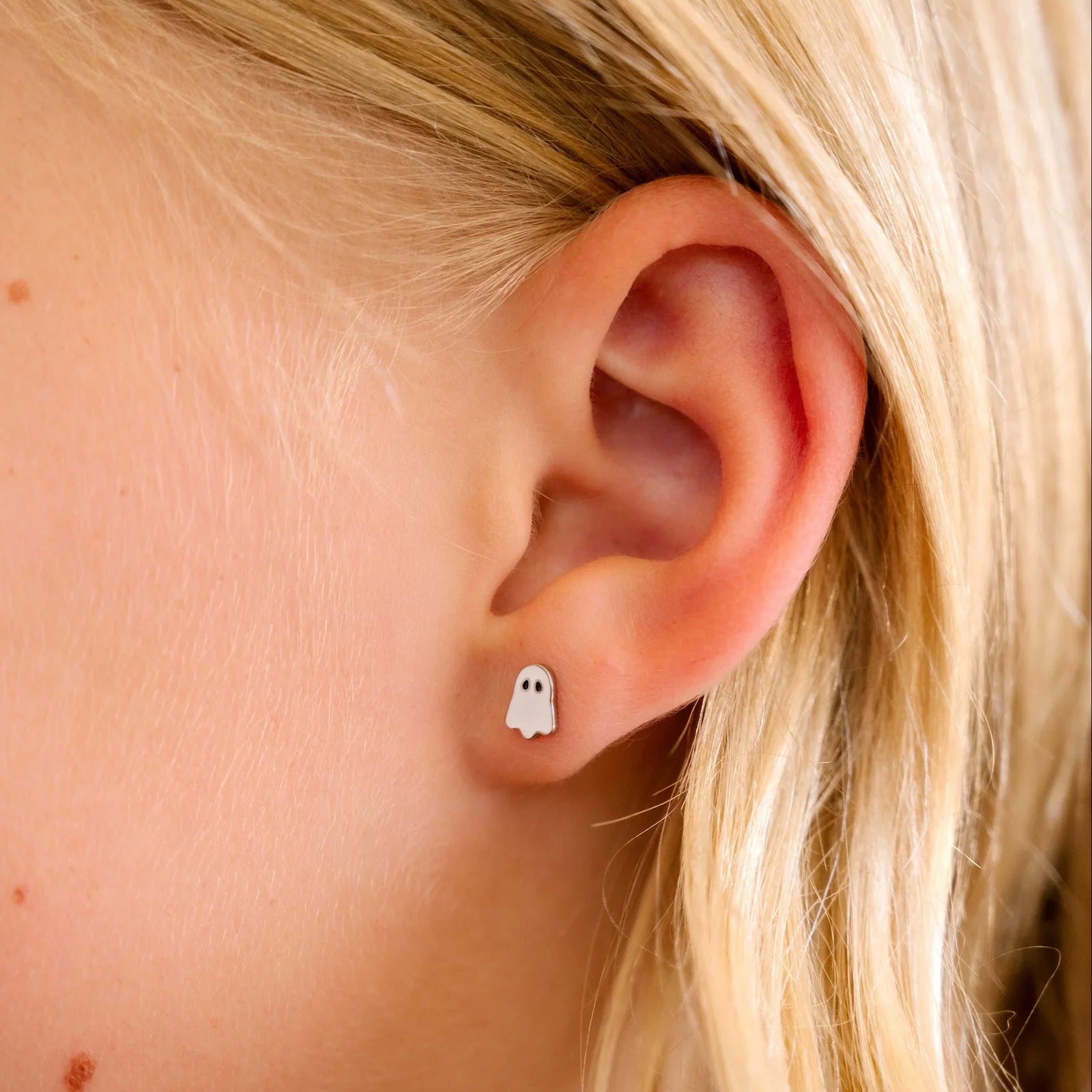 Close-up of an ear wearing a small ghost-shaped earring with blonde hair.