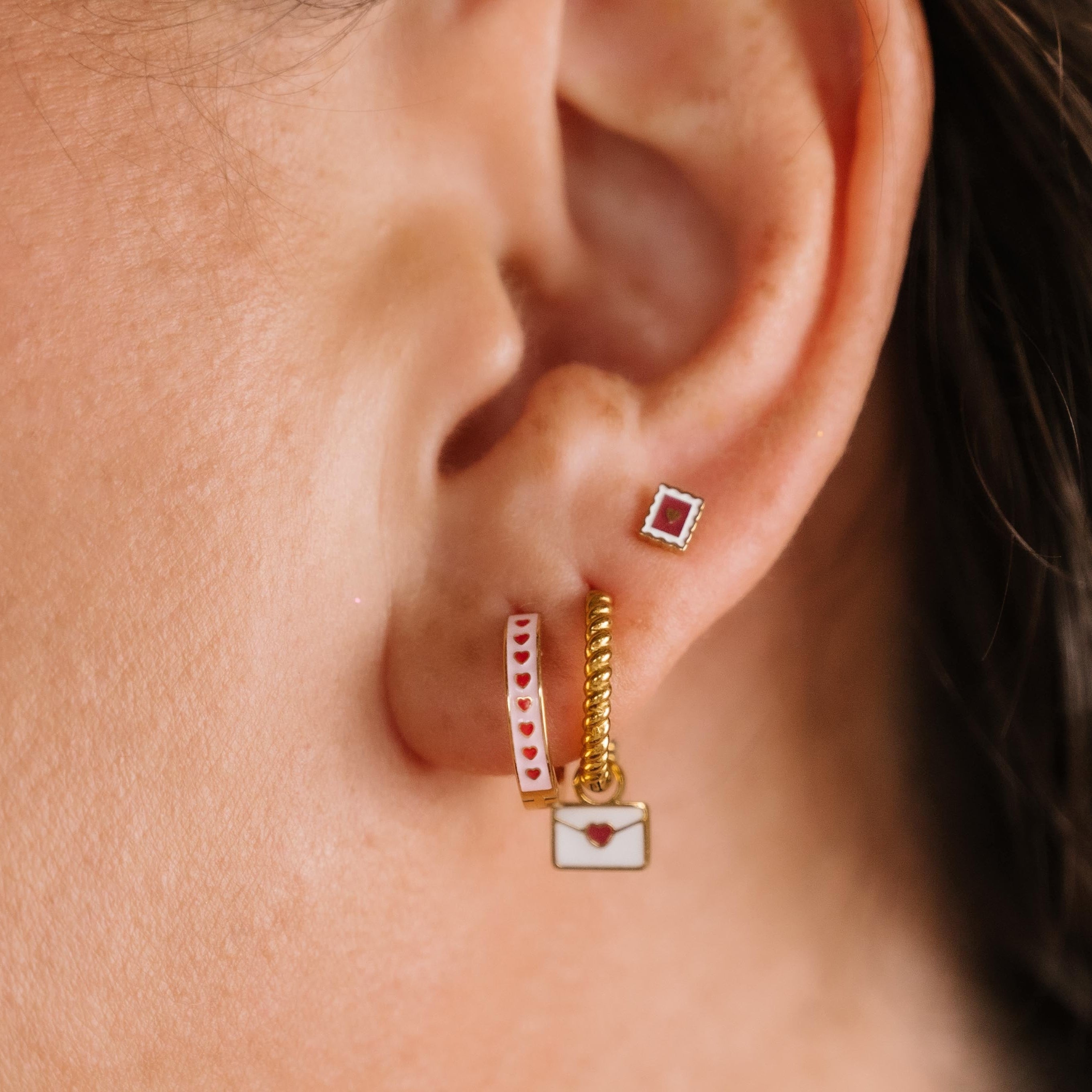 Close-up of an ear wearing gold and enamel hoop earrings with heart designs.