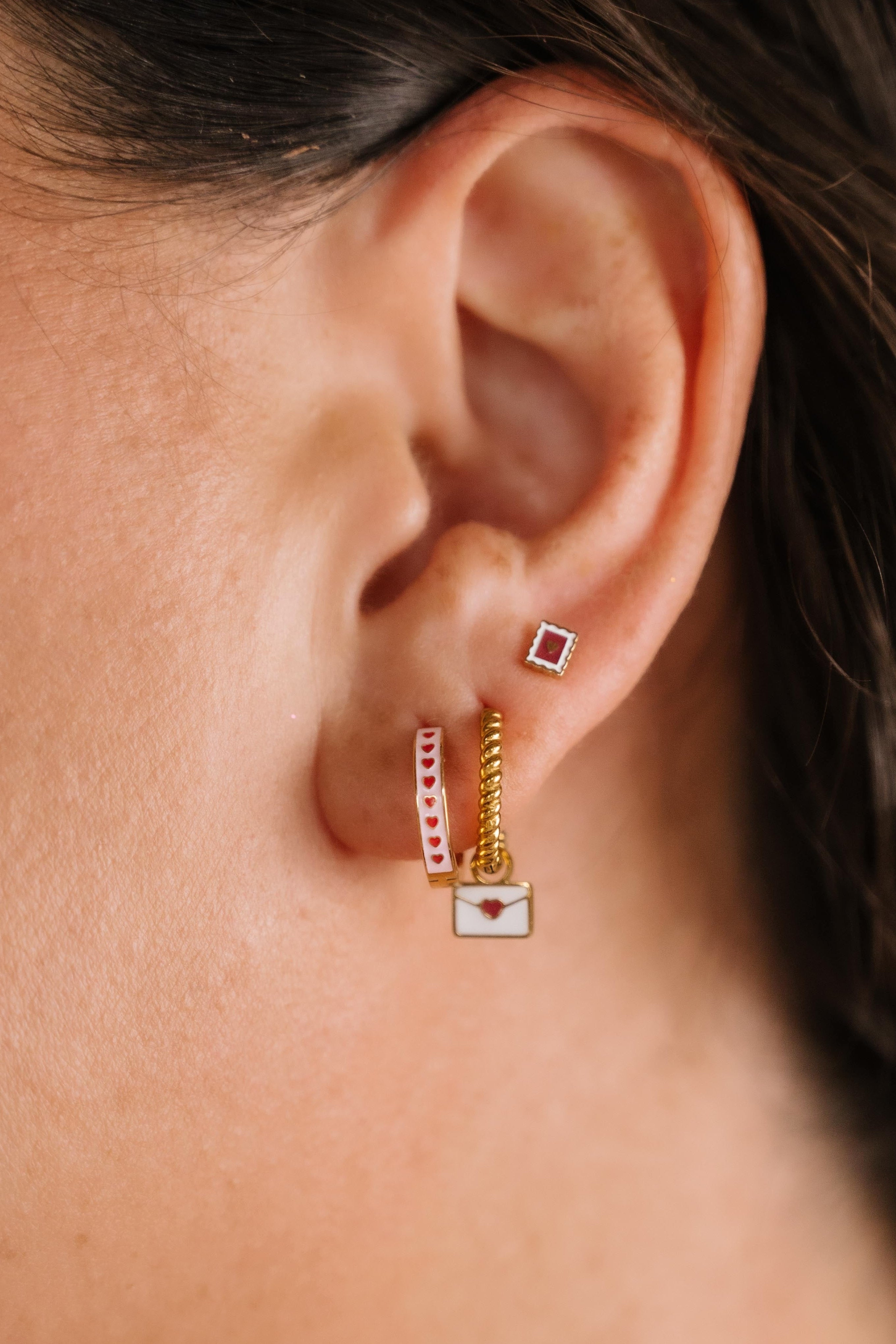 Close-up of an ear wearing gold and enamel hoop earrings with heart designs.