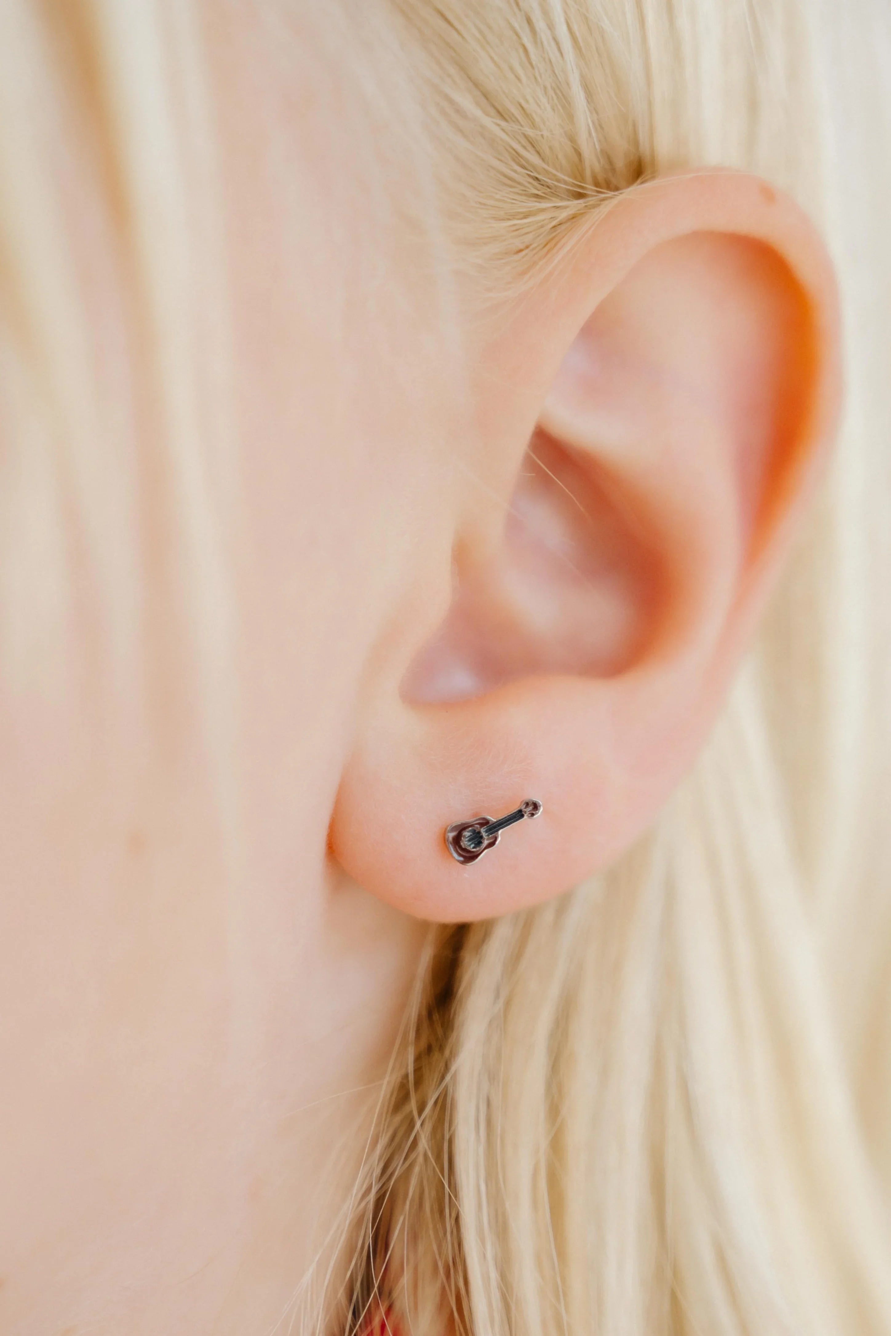 Close-up of an ear with a small brown guitar enamel stud earring against a blurred background.
