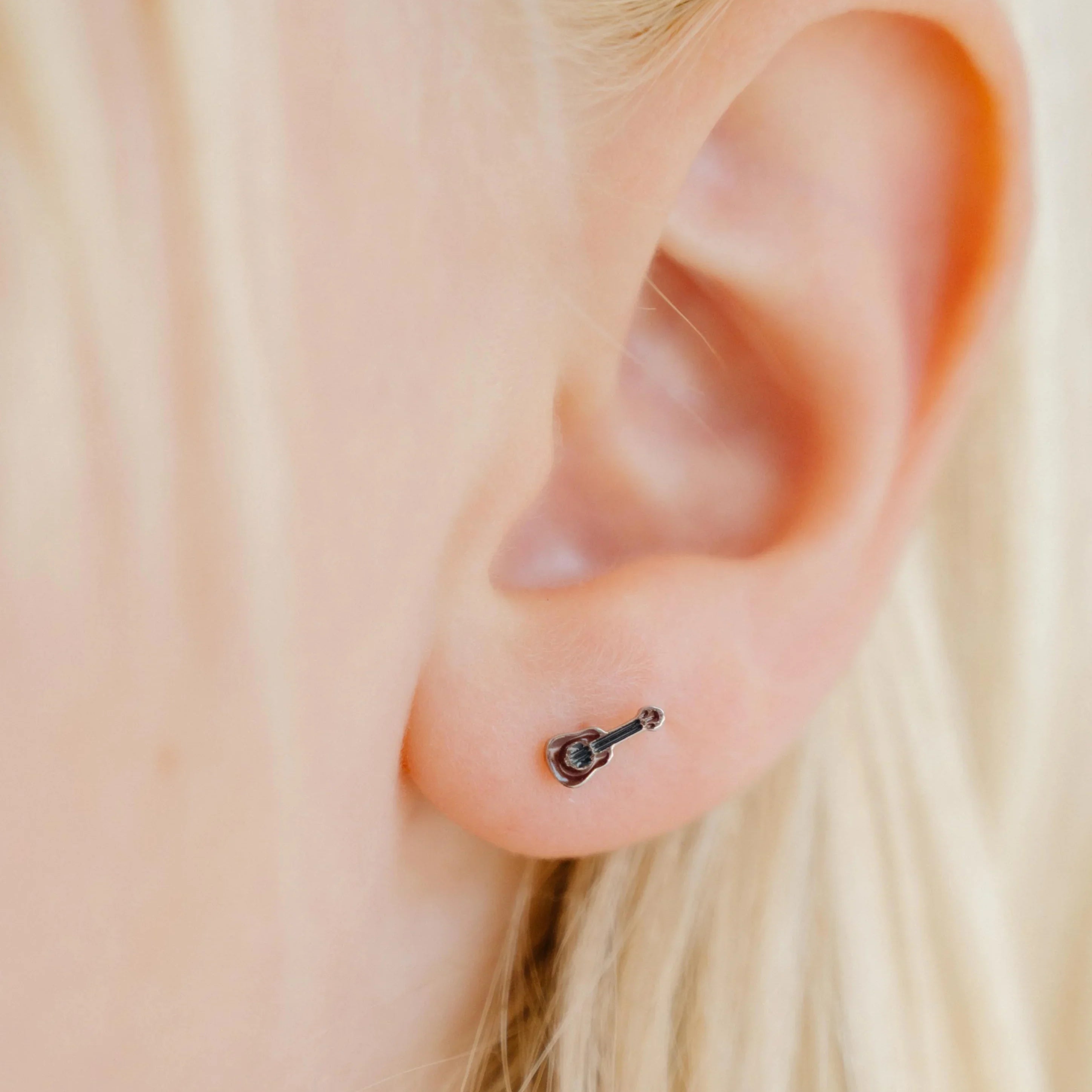 Close-up of an ear with a small brown guitar enamel stud earring against a blurred background.
