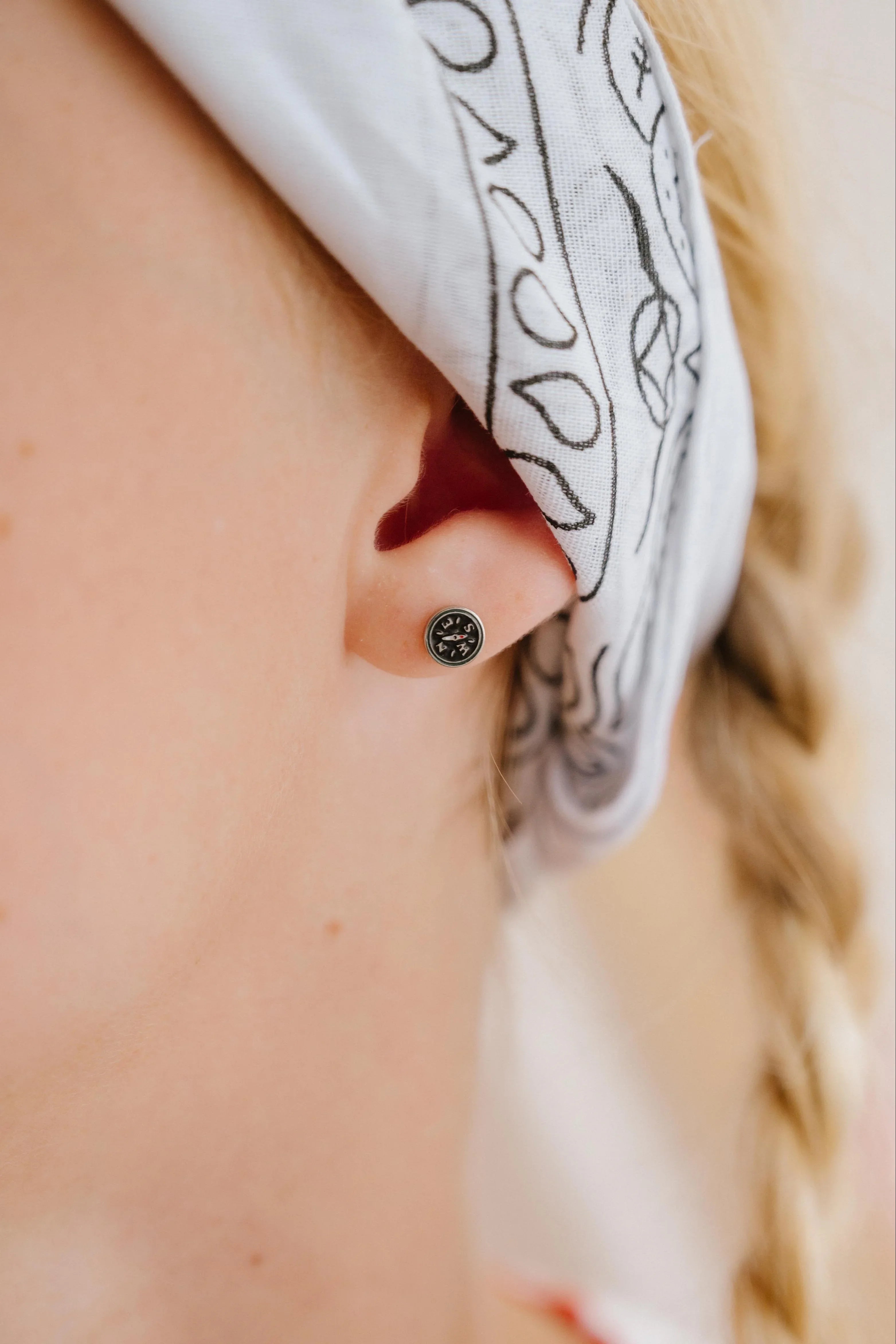 Close-up of person wearing a small compass-shaped enamel stud earring, wearing a white headscarf with black patterns.