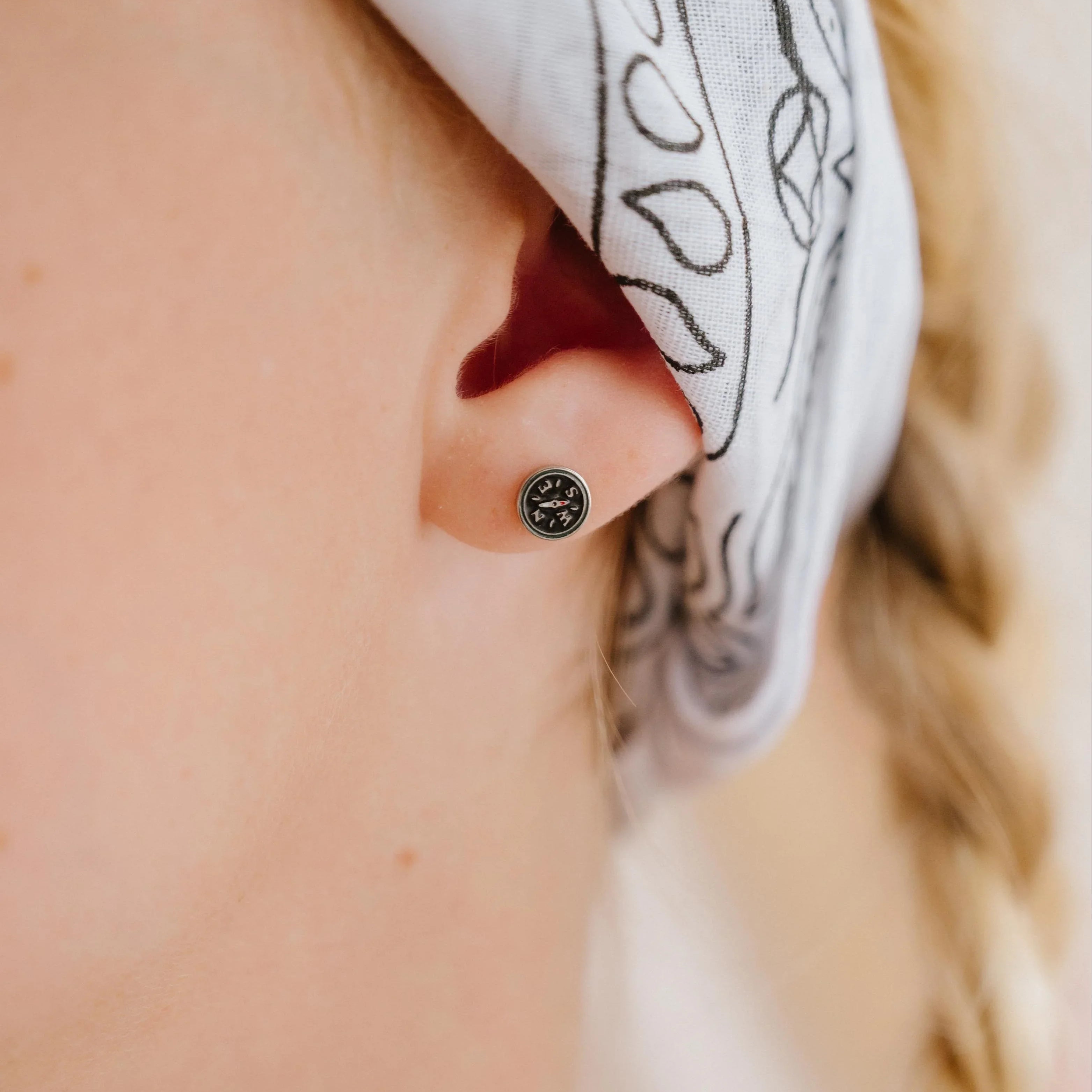 Close-up of person wearing a small compass-shaped enamel stud earring, wearing a white headscarf with black patterns.