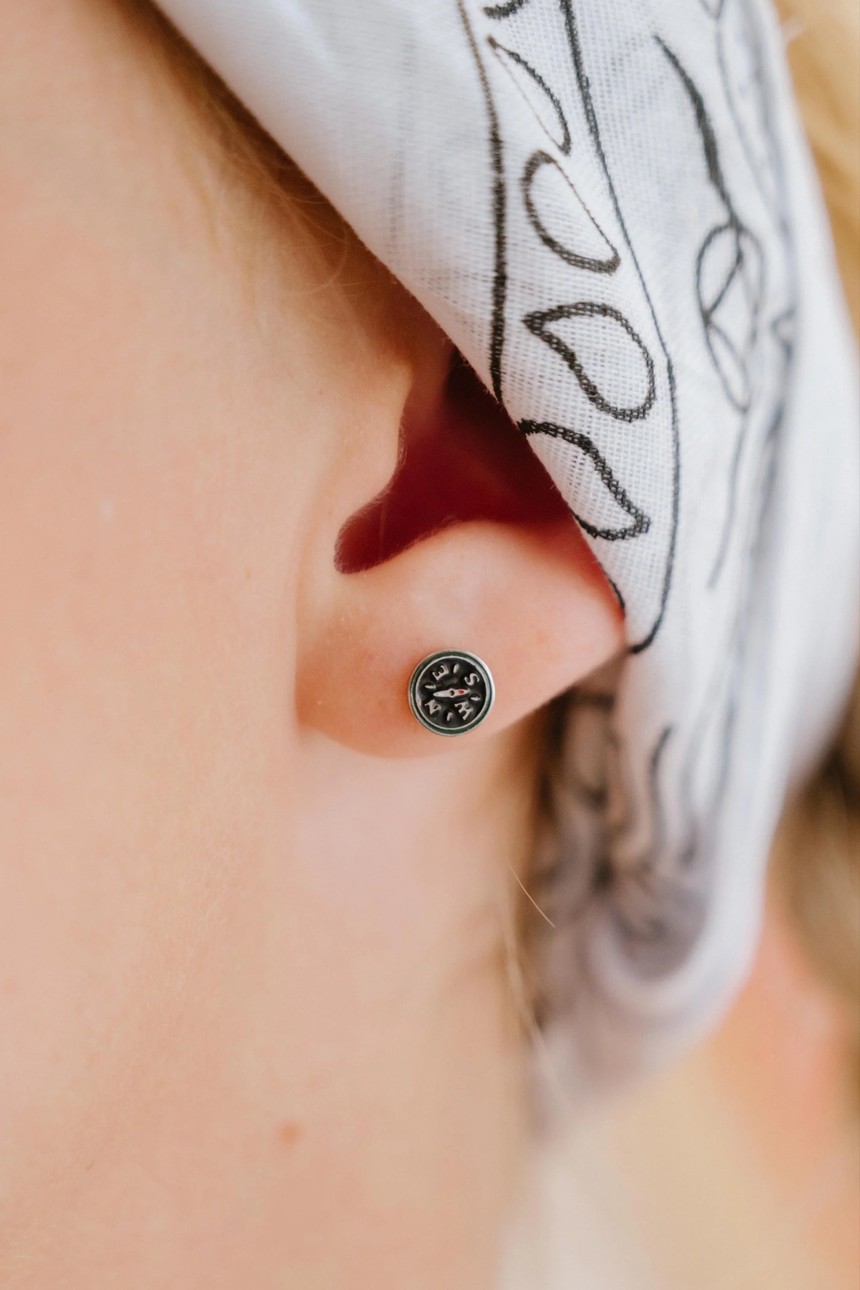 Close-up of an ear with a small compass-shaped enamel stud earring, wearing a white headscarf with black patterns.
