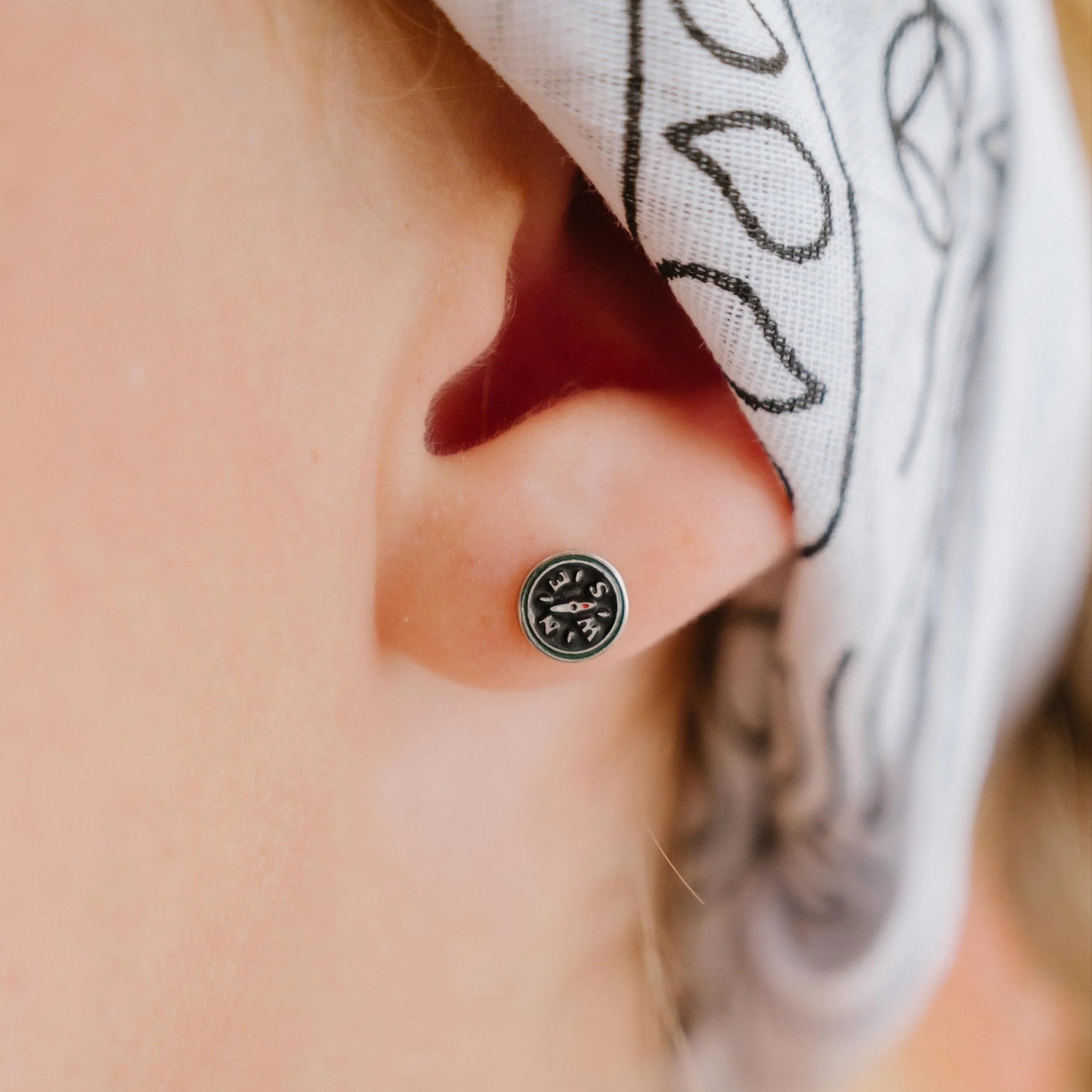Close-up of an ear with a small compass-shaped enamel stud earring, wearing a white headscarf with black patterns.