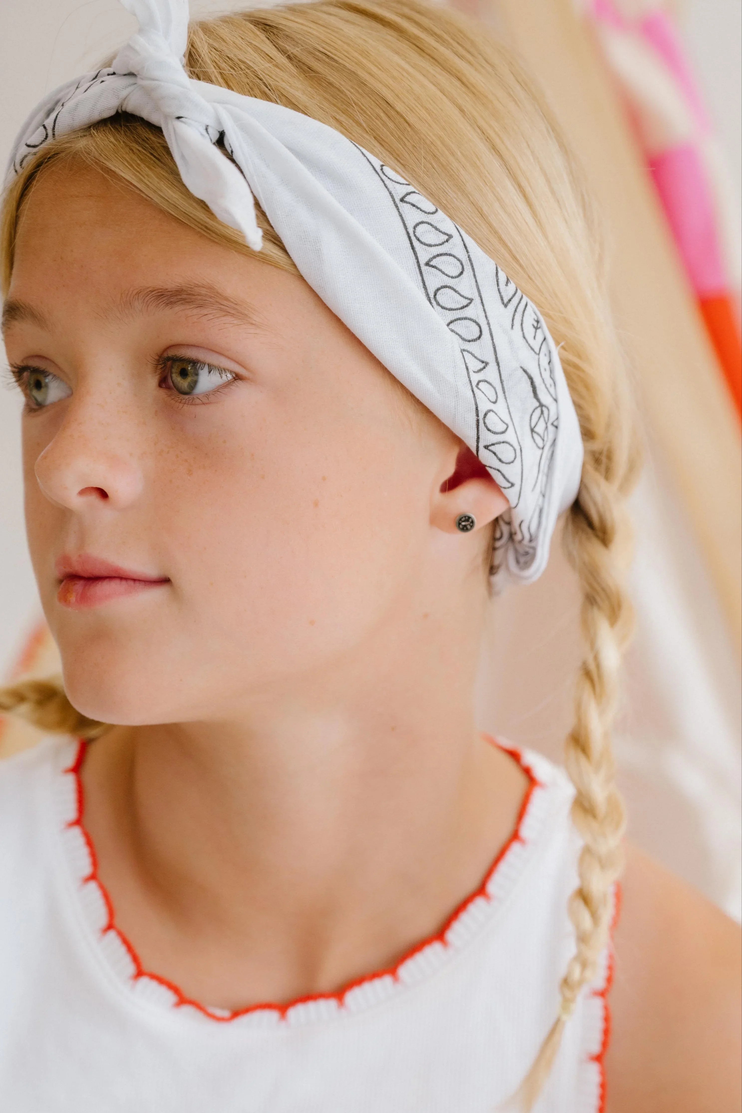 Young girl wearing a white tank top with red trim and cherry design, sitting indoors. Features a small enamel compass stud earring.