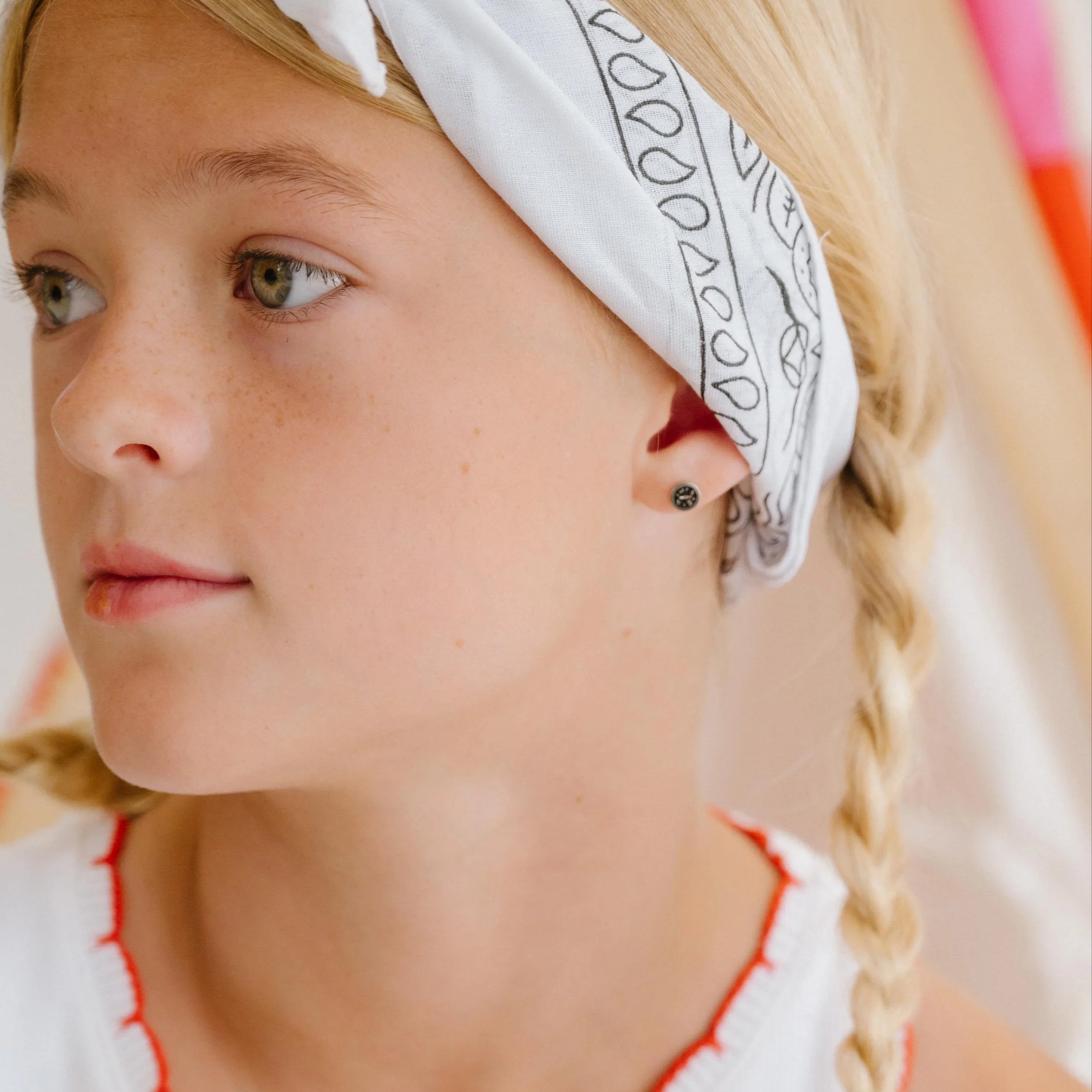 Young girl wearing a white tank top with red trim and cherry design, sitting indoors. Features a small enamel compass stud earring.