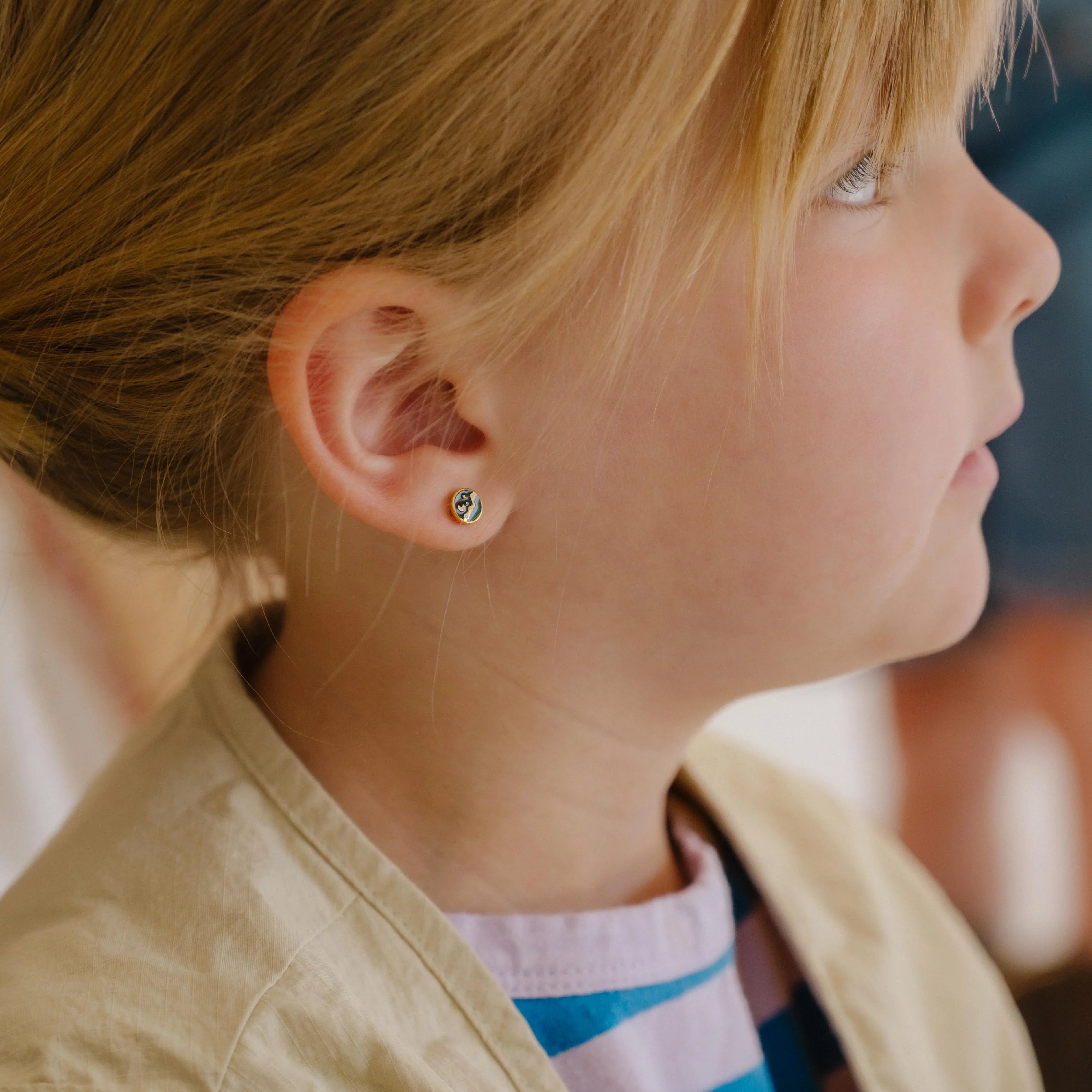 Close-up of child wearing a small circle enamel flat back stud earring that features a tiny night sky scene over a mountain landscape.