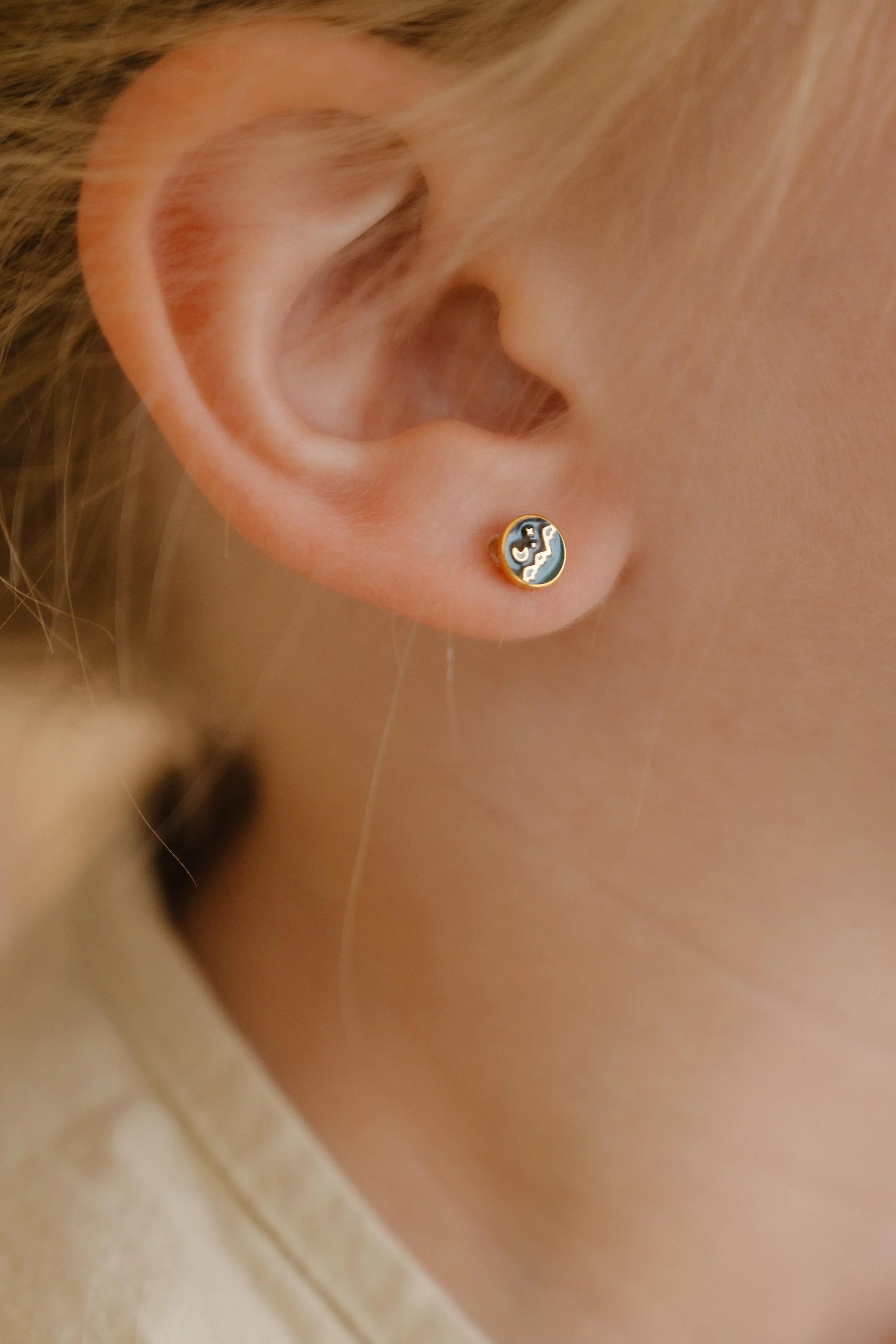 Close-up of an ear wearing a small circle enamel flat back stud earring that features a tiny night sky scene over a mountain landscape.