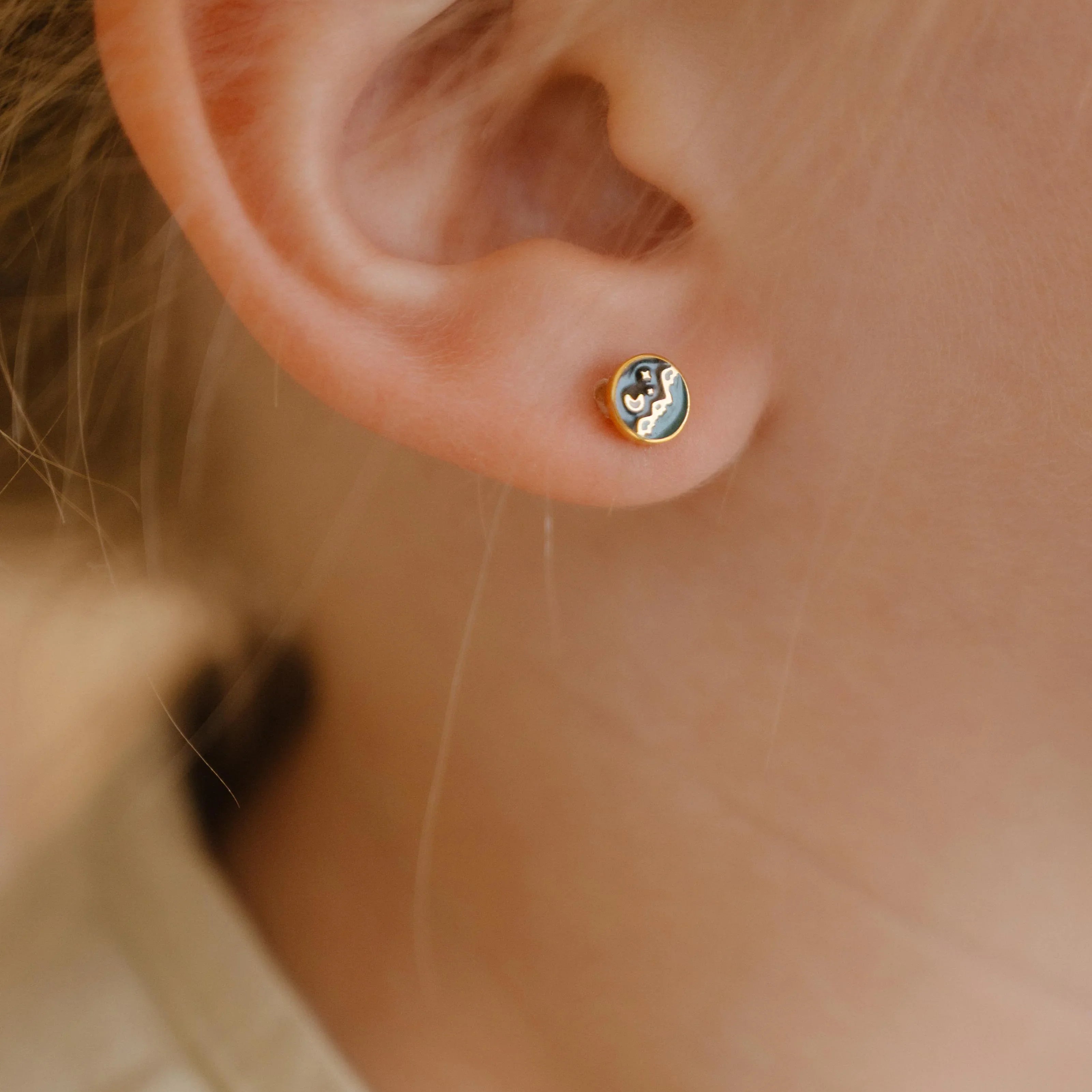 Close-up of an ear wearing a small circle enamel flat back stud earring that features a tiny night sky scene over a mountain landscape.