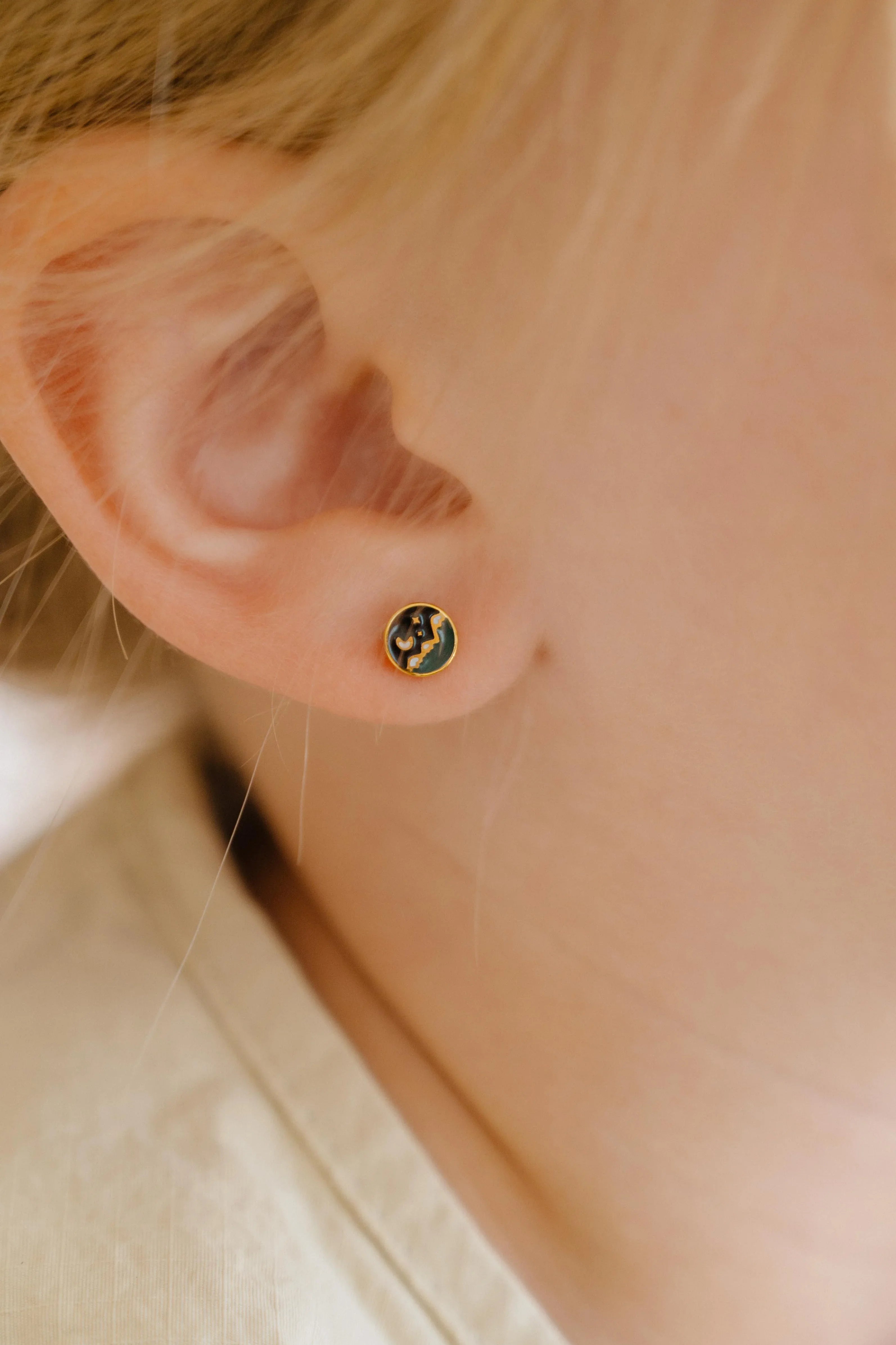 Close-up of an person wearing a small circle enamel flat back stud earring that features a tiny night sky scene over a mountain landscape.