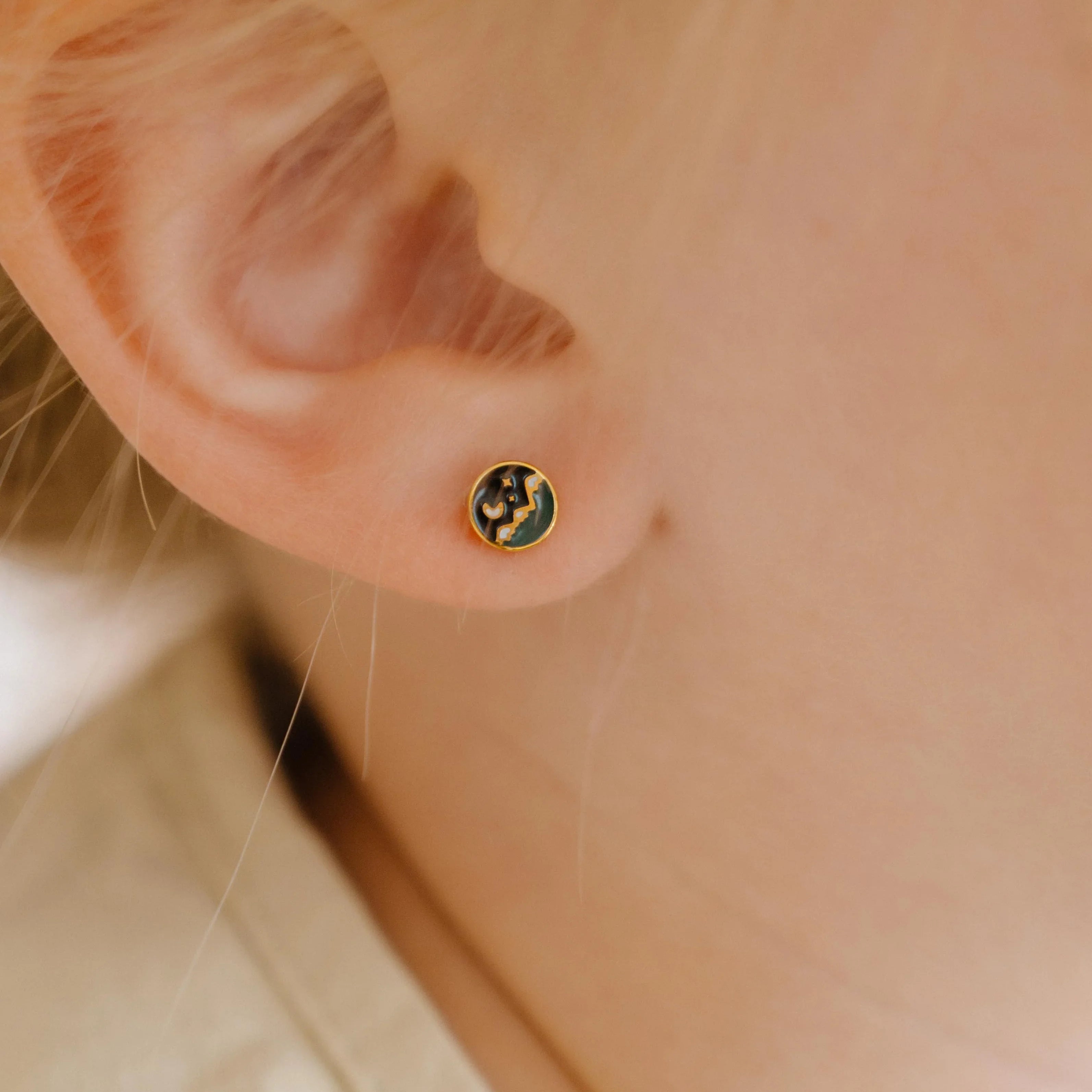 Close-up of an person wearing a small circle enamel flat back stud earring that features a tiny night sky scene over a mountain landscape.