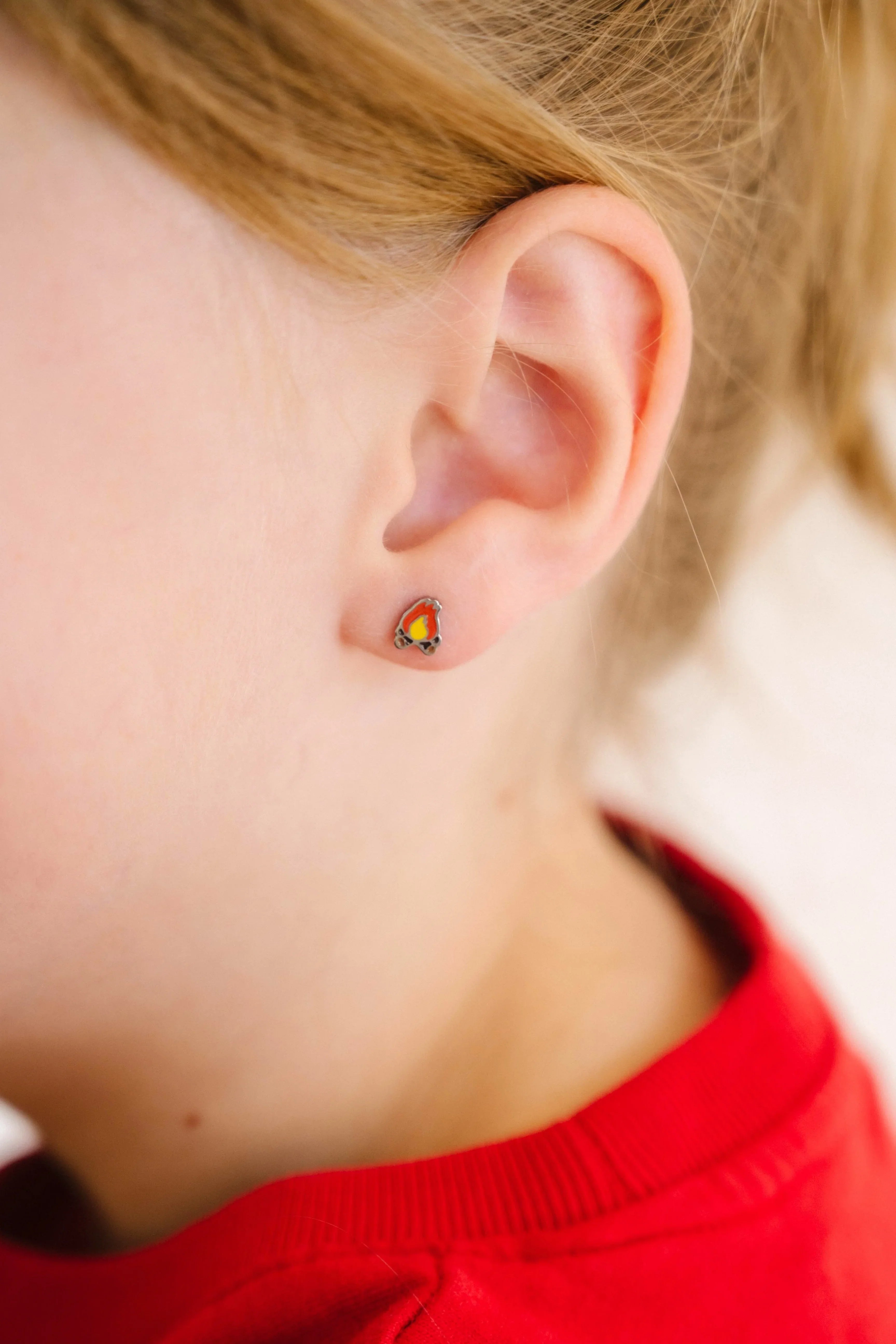 Close-up of a child's ear wearing a small, colorful enamel campfire design stud earring.
