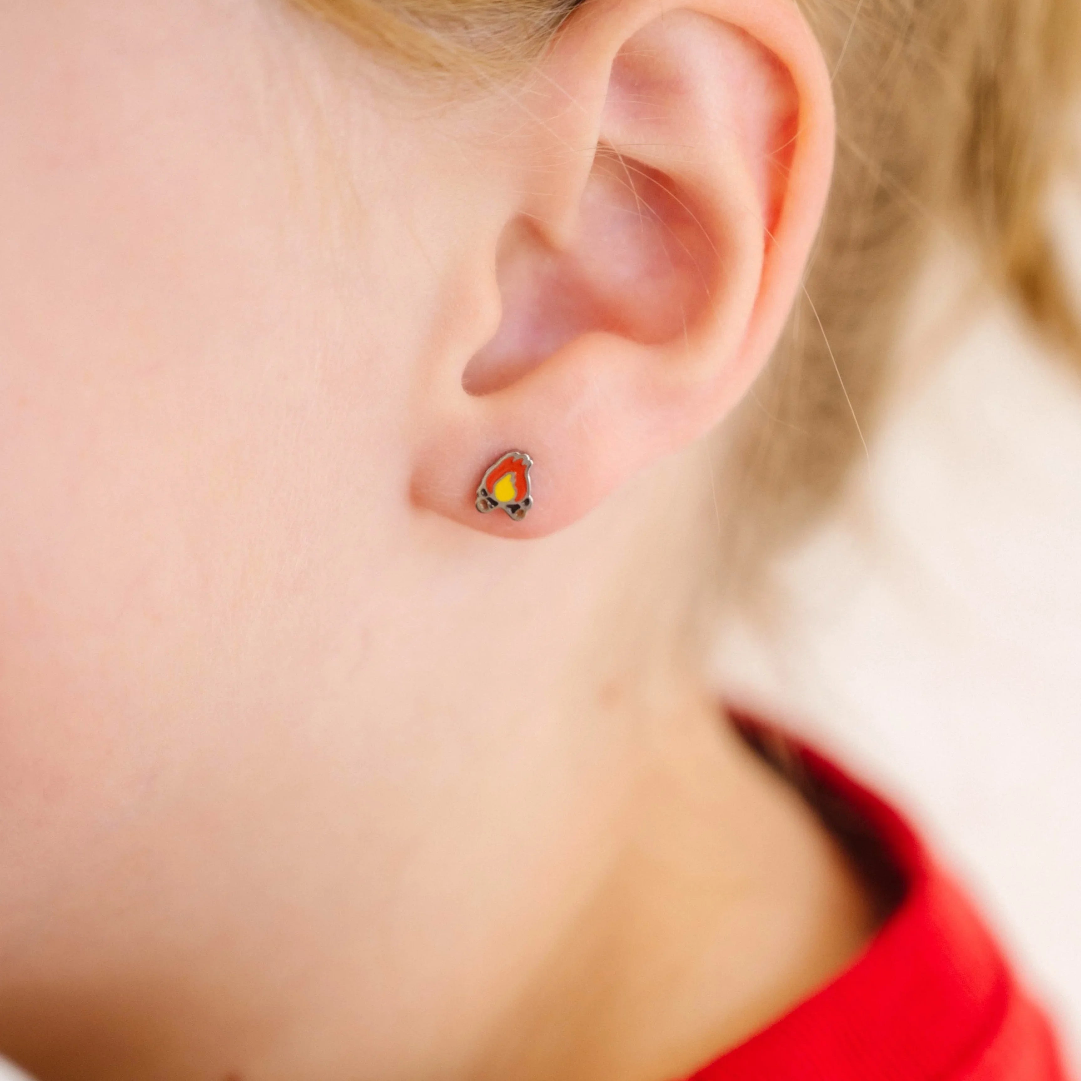 Close-up of a child's ear wearing a small, colorful enamel campfire design stud earring.