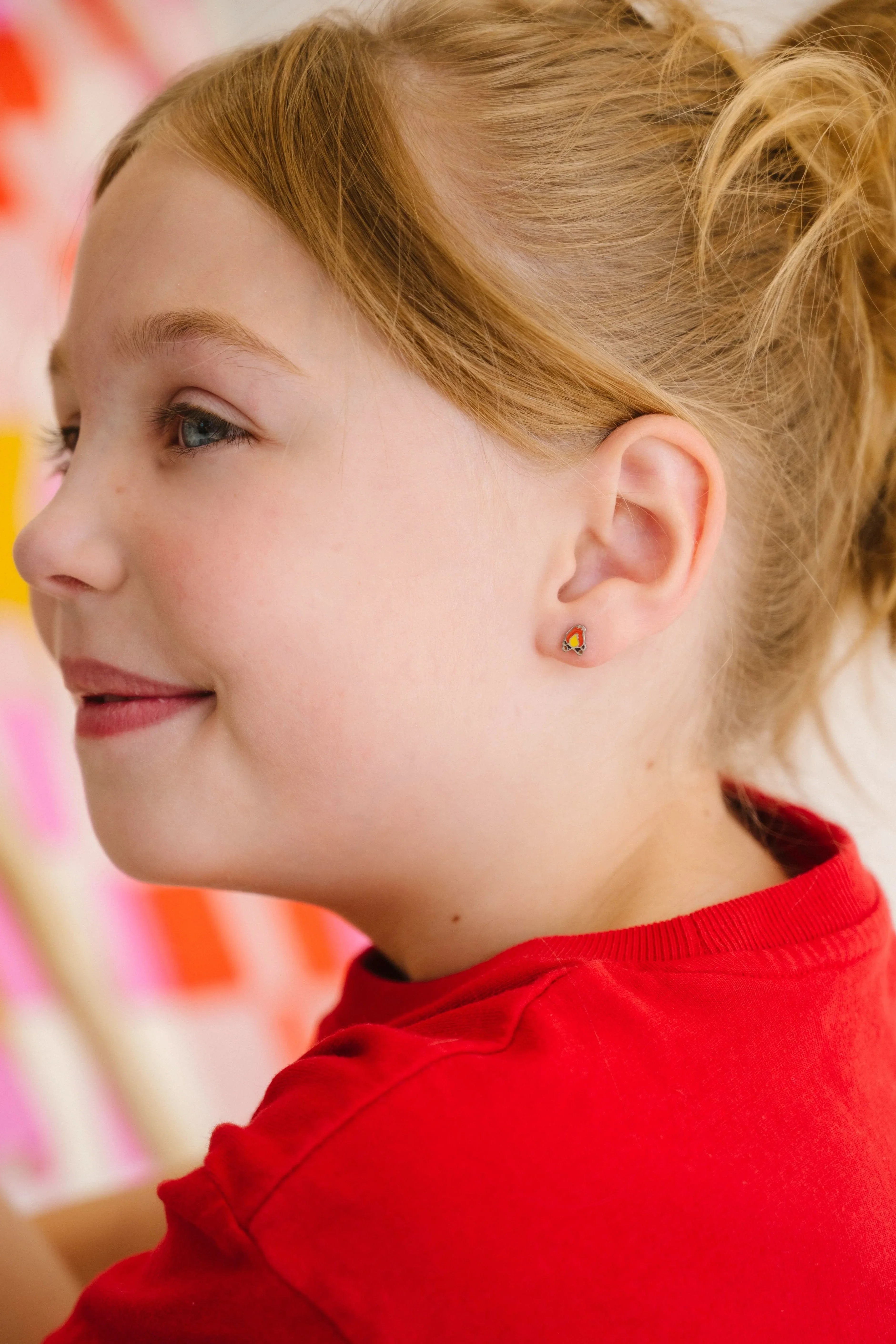 Close-up of a smiling child in a red shirt, with a ponytail, wearing a small, colorful enamel campfire design stud earring.