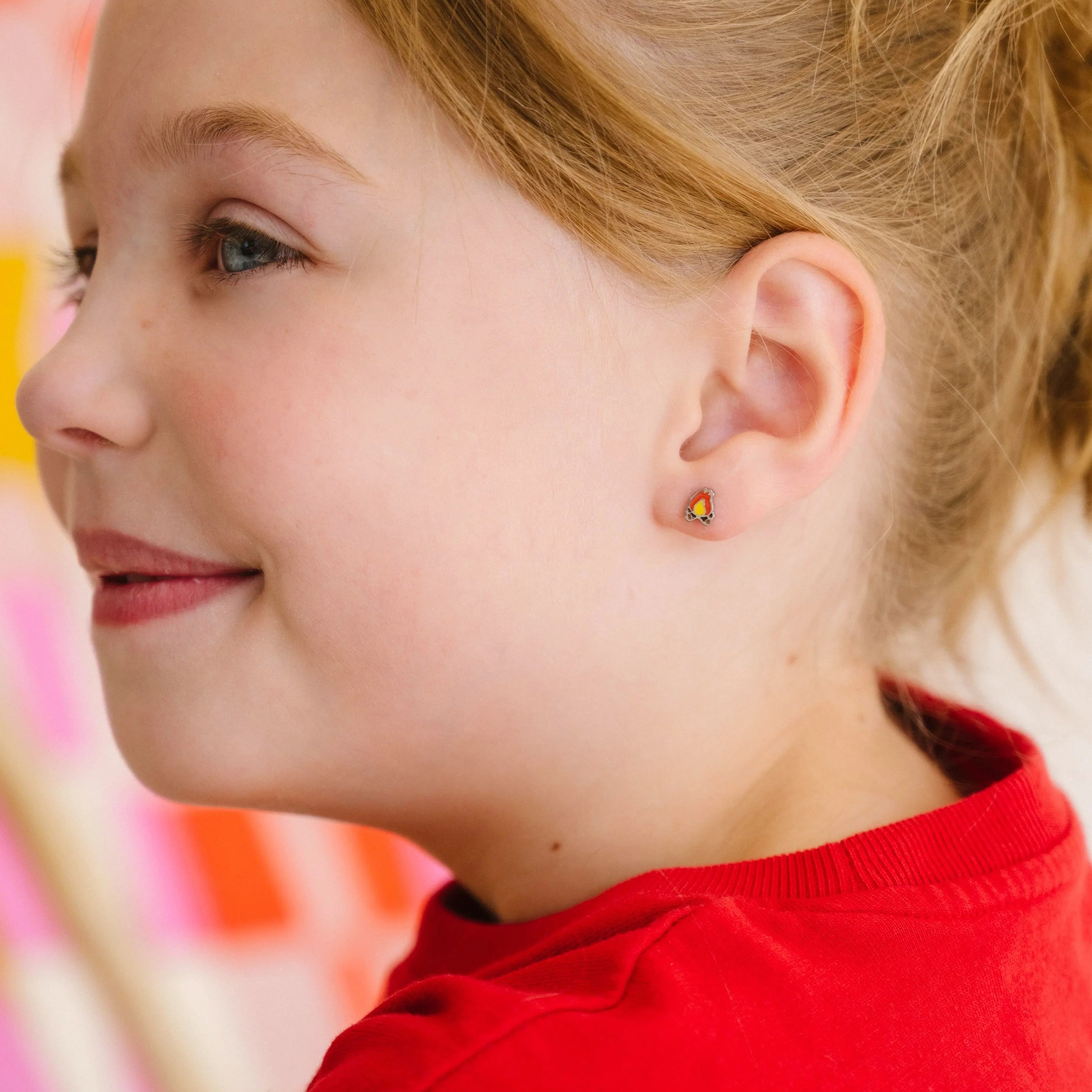 Close-up of a smiling child in a red shirt, with a ponytail, wearing a small, colorful enamel campfire design stud earring.