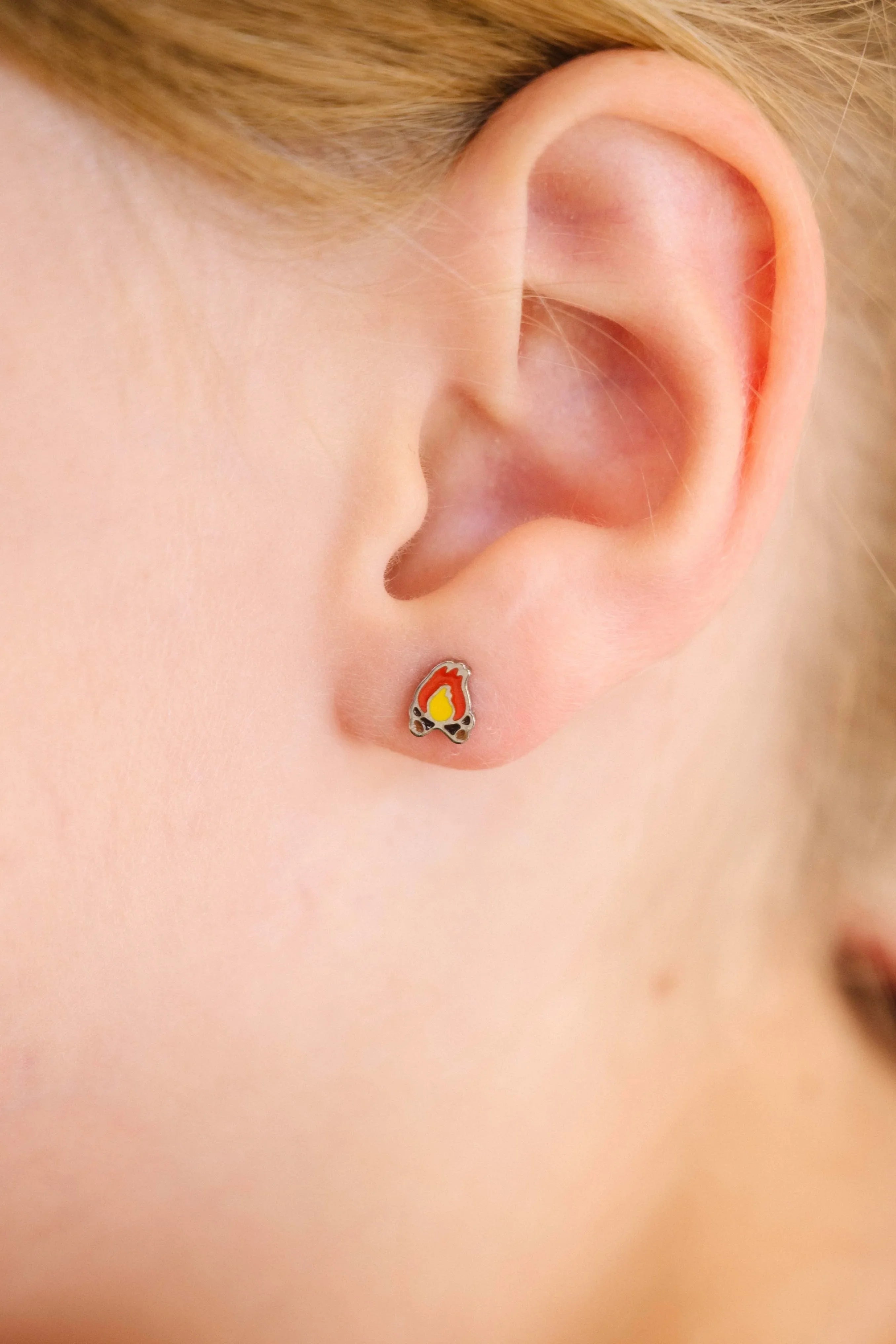 Close-up of a child's ear wearing a small, colorful enamel campfire design stud earring.