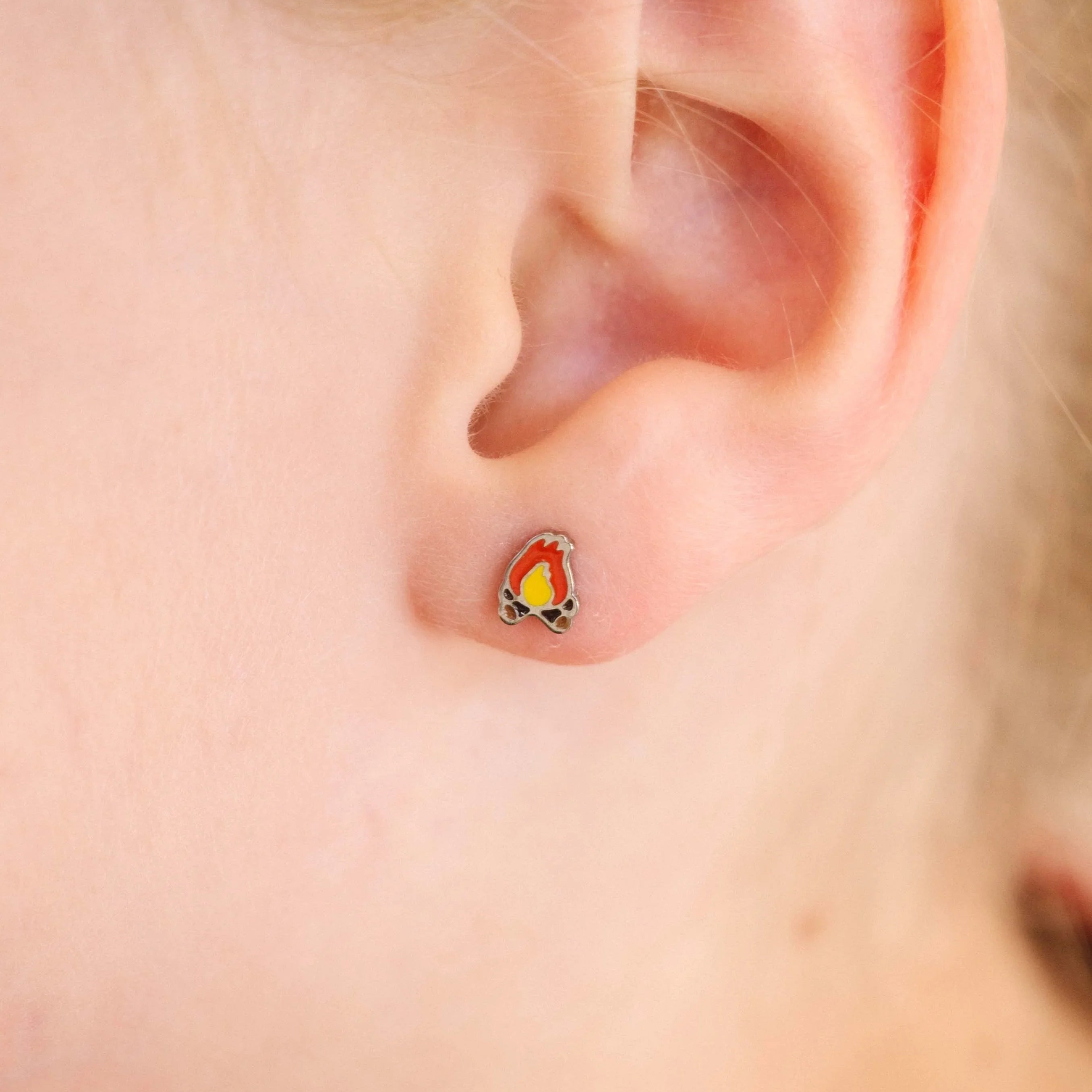 Close-up of a child's ear wearing a small, colorful enamel campfire design stud earring.