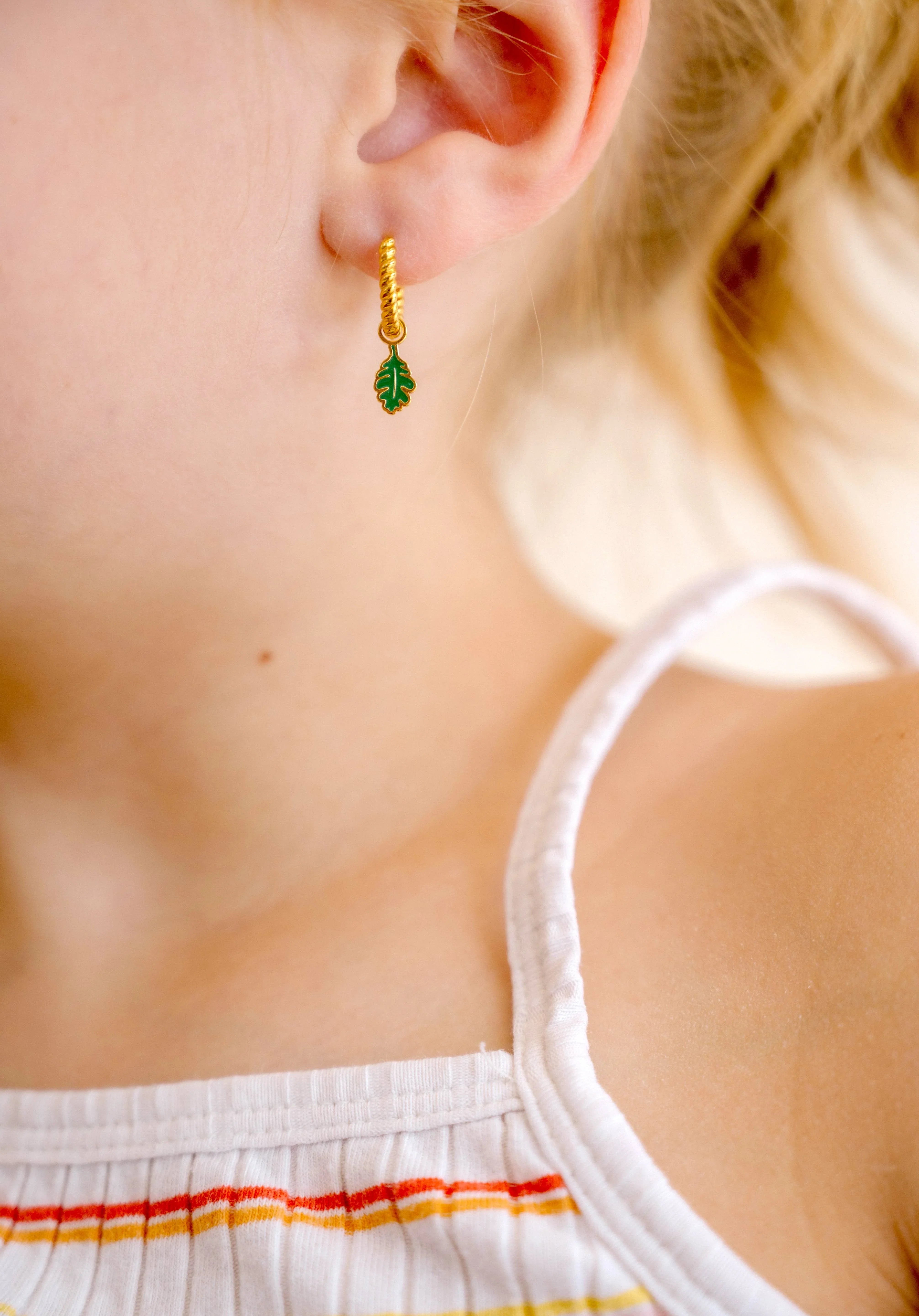 Close-up of a child wearing gold hoop earrings with green and gold oak leaf enamel hoop charm.